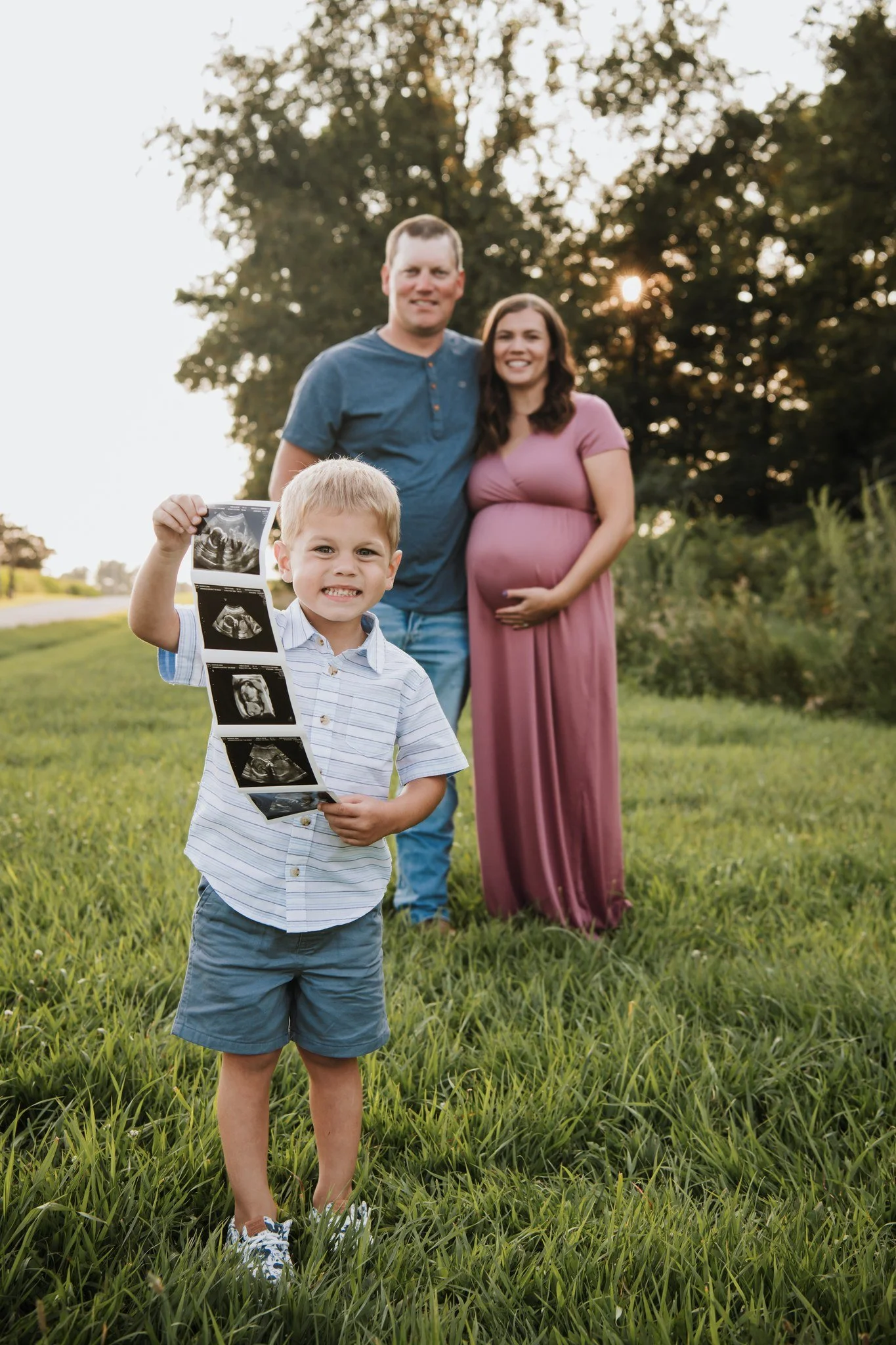 A young boy holds up an ultrasound scan while standing on a grassy field with happy expecting parents in the background, during sunset.