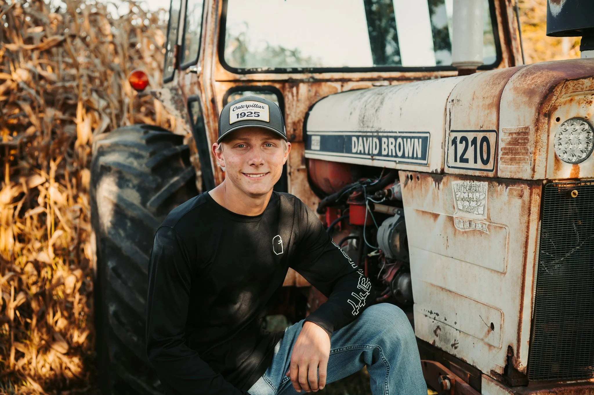 A young man dressed in a black long-sleeve shirt and blue jeans, wearing a cap that says 'Caterpillar 1925,' sitting next to an old, rusted tractor with the name 'David Brown' on it, outdoors in a field of dried corn stalks.