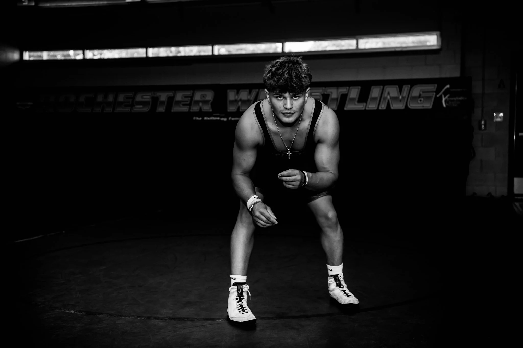 A wrestler in athletic attire crouching in a wrestling ring, with a sign reading 'Rochester Wrestling' in the background.