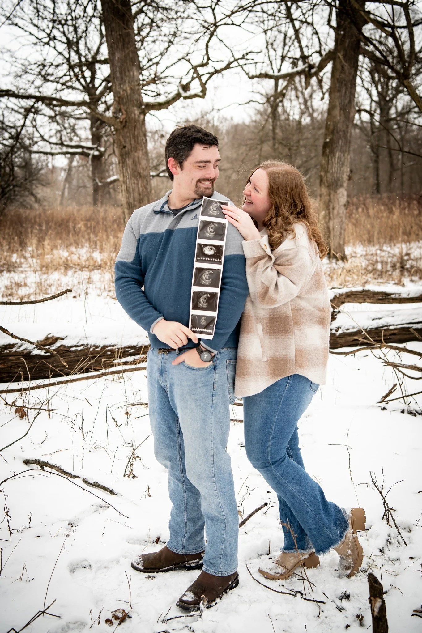 Couple standing outdoors in snow, holding an ultrasound photo, smiling at each other, in a winter forest setting.