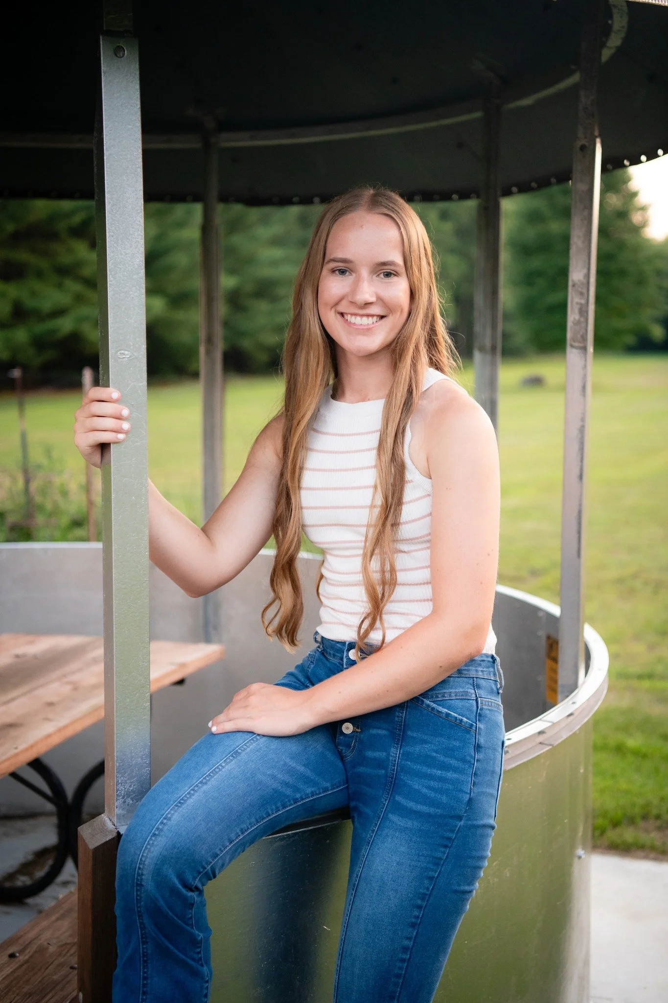 A young woman with long reddish-brown hair, smiling, sitting on a metal and wood structure in outdoor park setting with green trees in background.