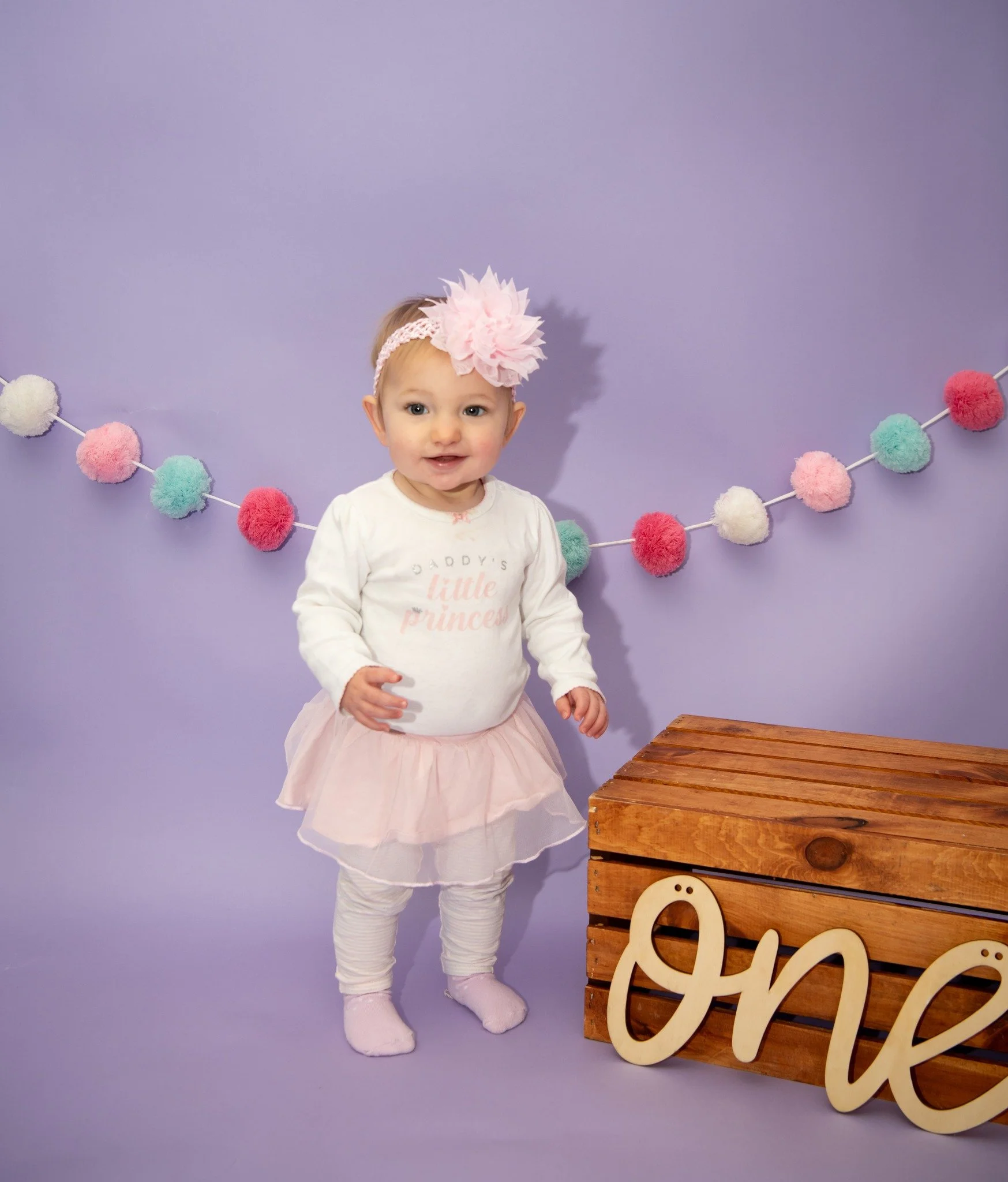 A baby girl standing indoors against a purple background decorated with colorful pom-pom garland. She wears a white long-sleeve shirt with pink text, pink tutu skirt, white leggings, pink socks, and a pink headband with a large flower. There is a woo