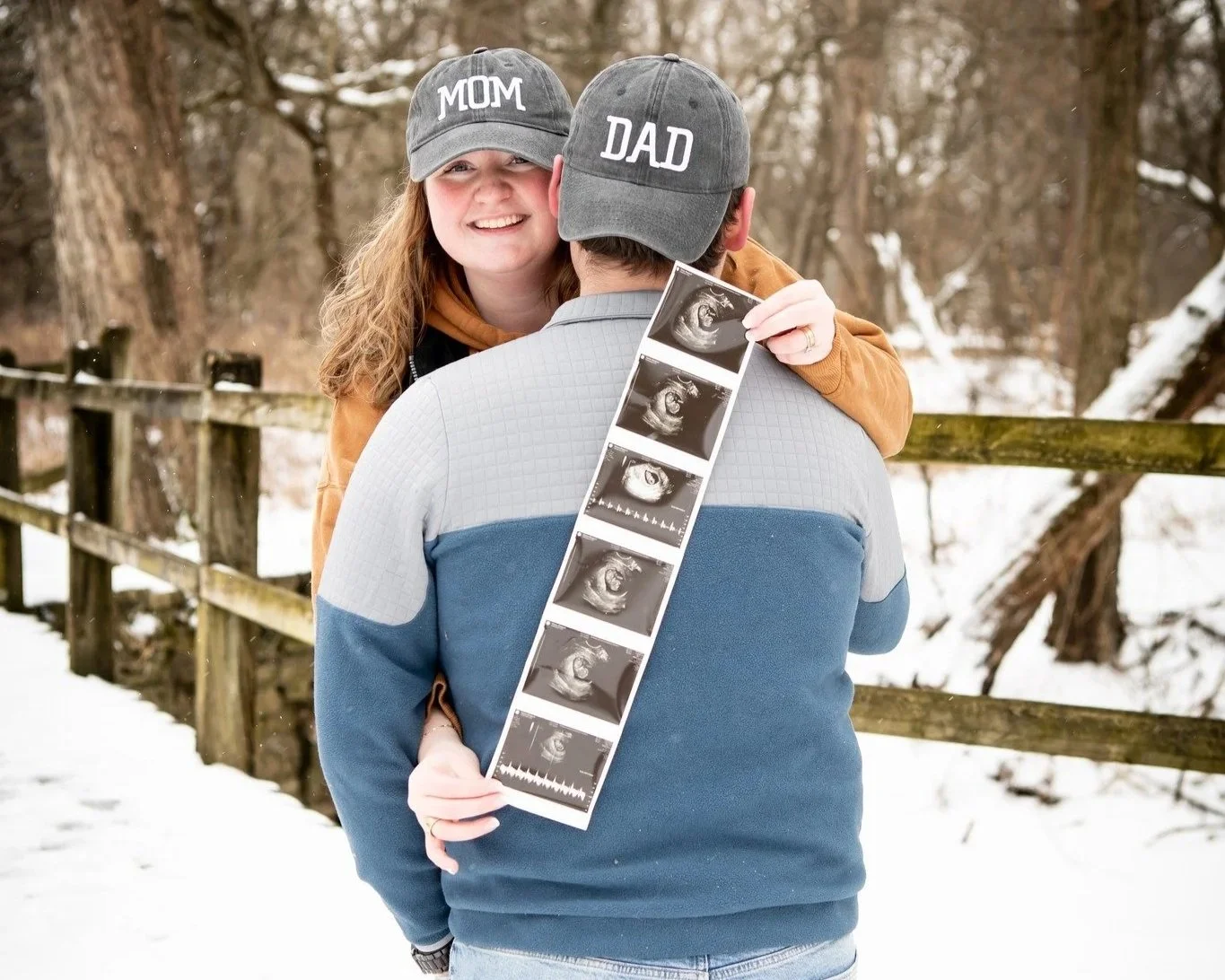 A woman and man wearing matching hats labeled 'MOM' and 'DAD' embrace in a snowy outdoor setting, with the woman smiling and holding ultrasound images of a baby in front of the man's back.