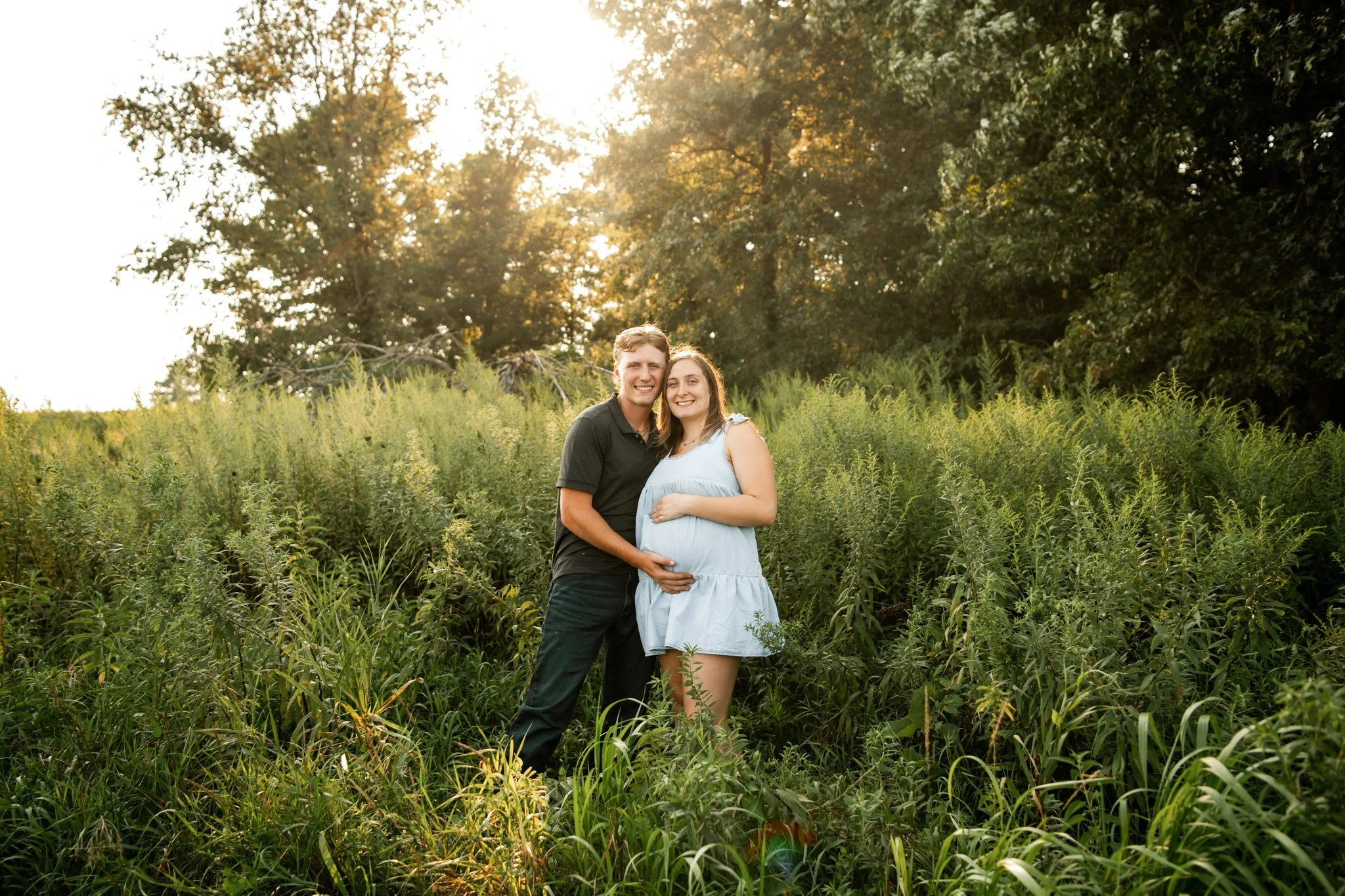 A happy pregnant woman and man standing together in a lush green field during sunset, smiling and holding her belly.