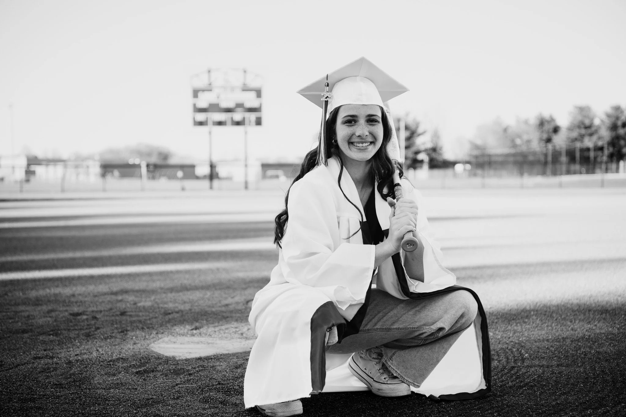 Young woman in graduation cap and gown crouching on a football field, smiling and holding a diploma, with a scoreboard and trees in the background.