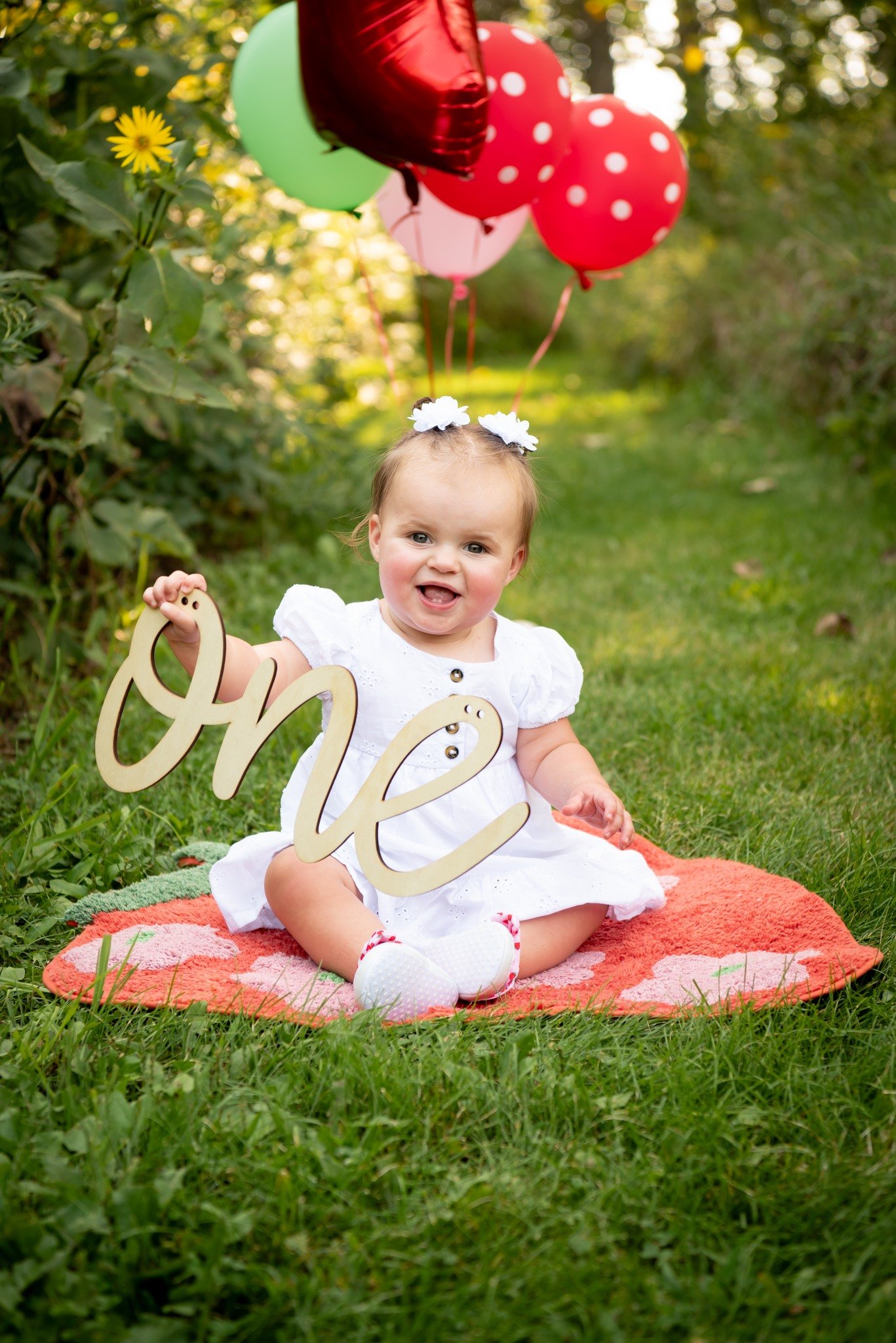 A young girl sitting on a pink blanket outdoors, holding a wooden sign that says 'one', with balloons behind her, wearing a white dress and white shoes, celebrating her first birthday.