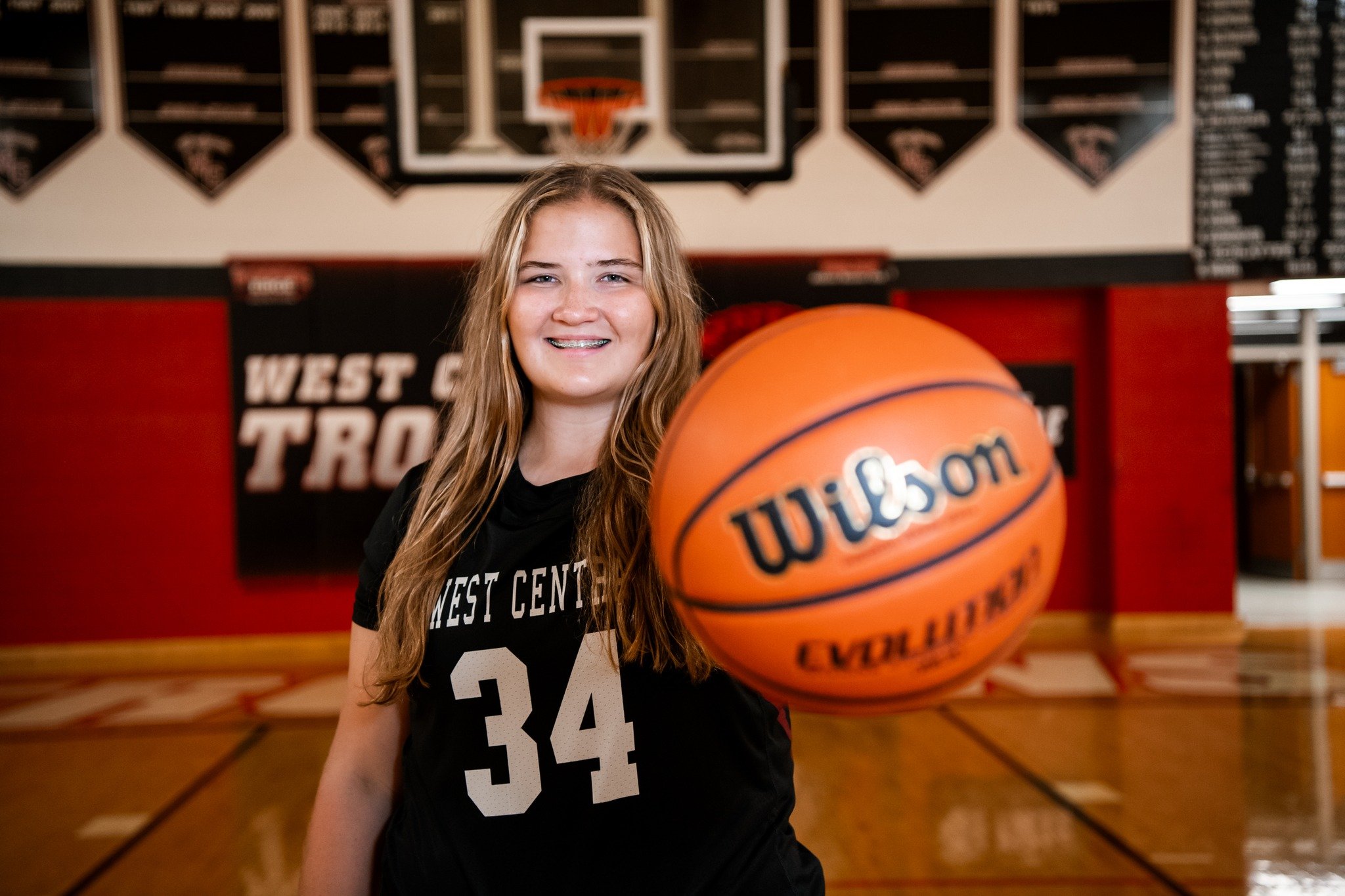 A young female basketball player standing on an indoor court, holding a Wilson basketball towards the camera. She is wearing a black jersey with the number 34 and smiling. In the background, there are basketball hoops and banners.
