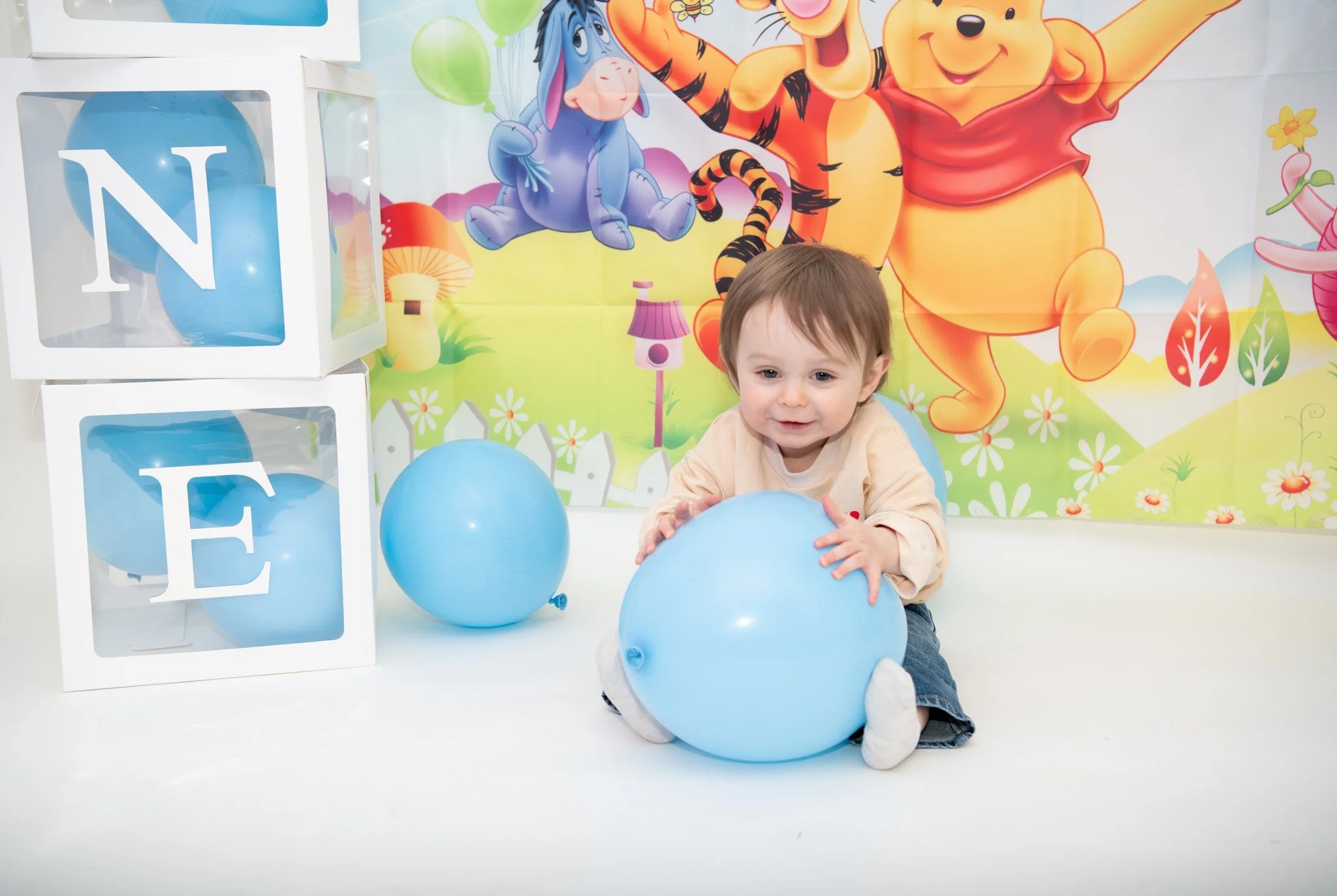 Young child sitting on the floor holding a blue balloon, with two additional blue balloons nearby, in front of a colorful backdrop featuring Winnie the Pooh, Tigger, and Eeyore characters, and white blocks with letters N and E in clear plastic window