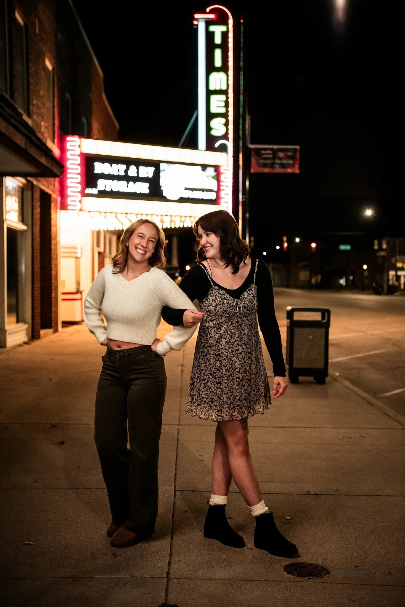 Two women walking on a sidewalk at night, smiling and holding onto each other's arms, with a neon-lit sign for a theater in the background.