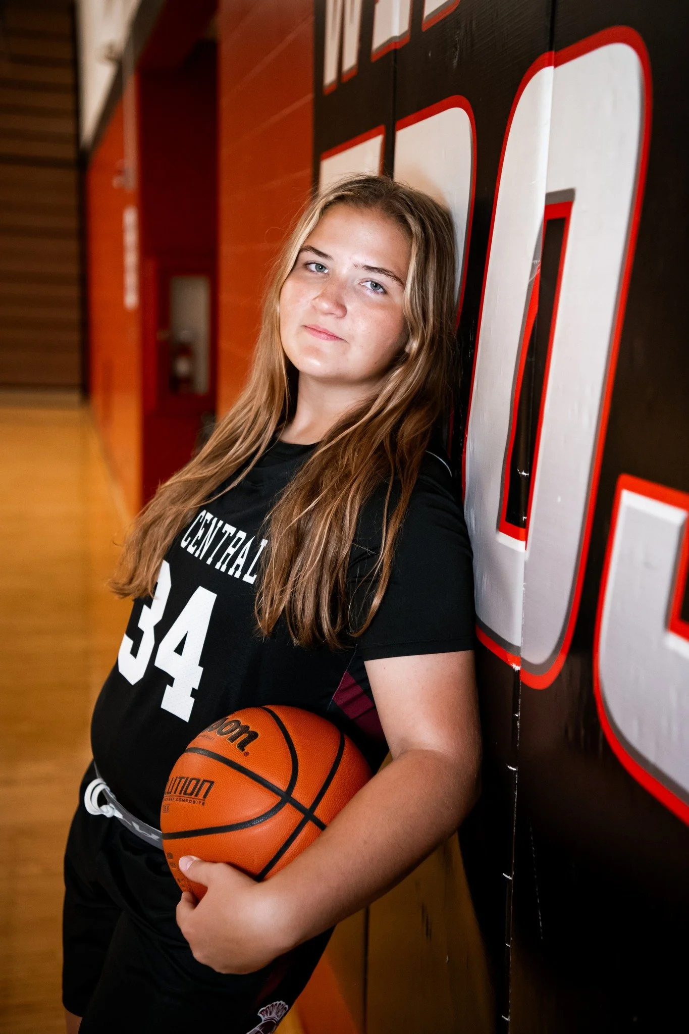 A teenage girl in a black sports jersey holding a basketball, leaning against a wall with large red and white lettering.