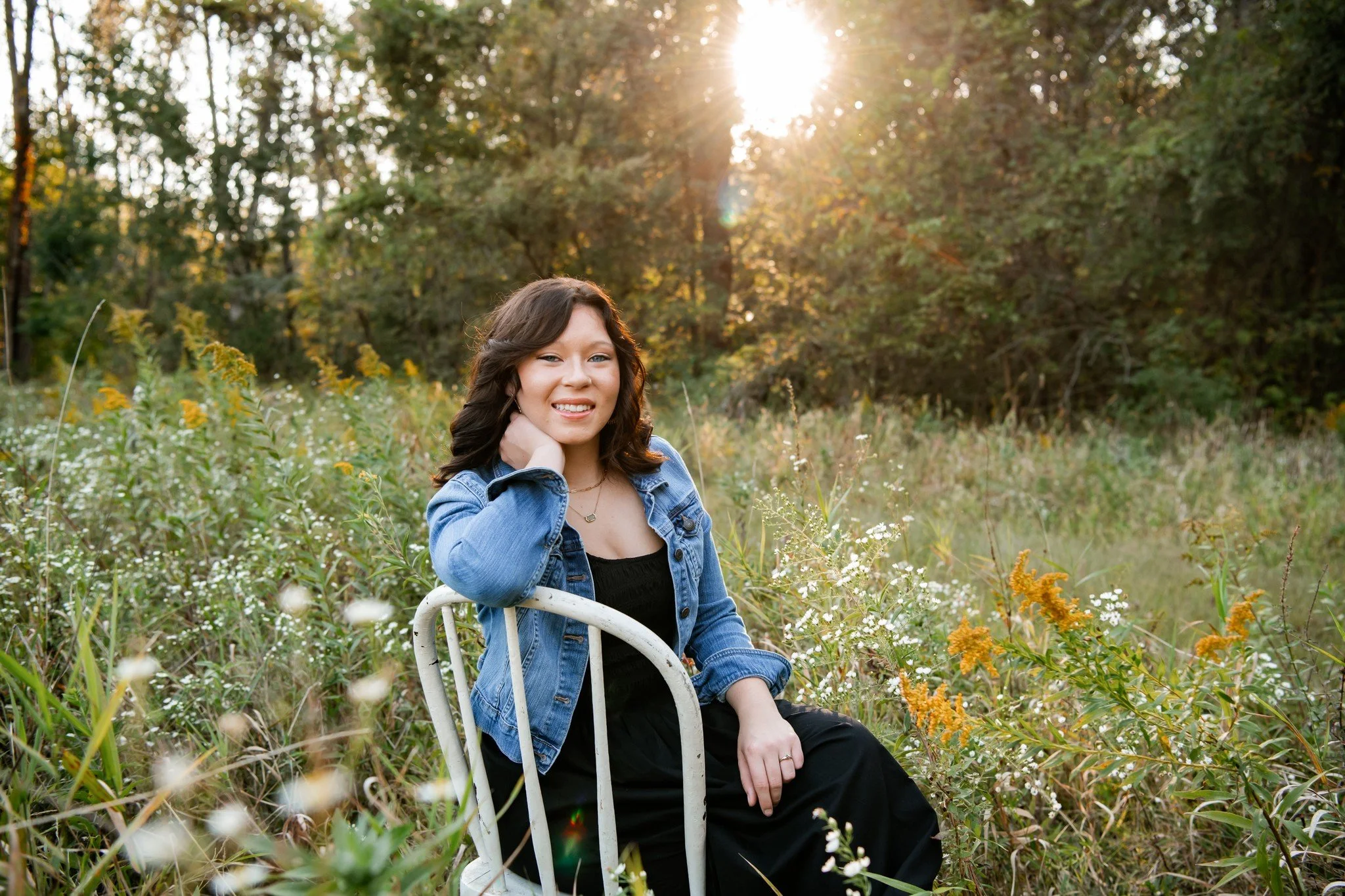 A young woman with wavy brown hair sitting on a white chair in a field of wildflowers, wearing a denim jacket and black dress, with the sun setting behind trees in the background.
