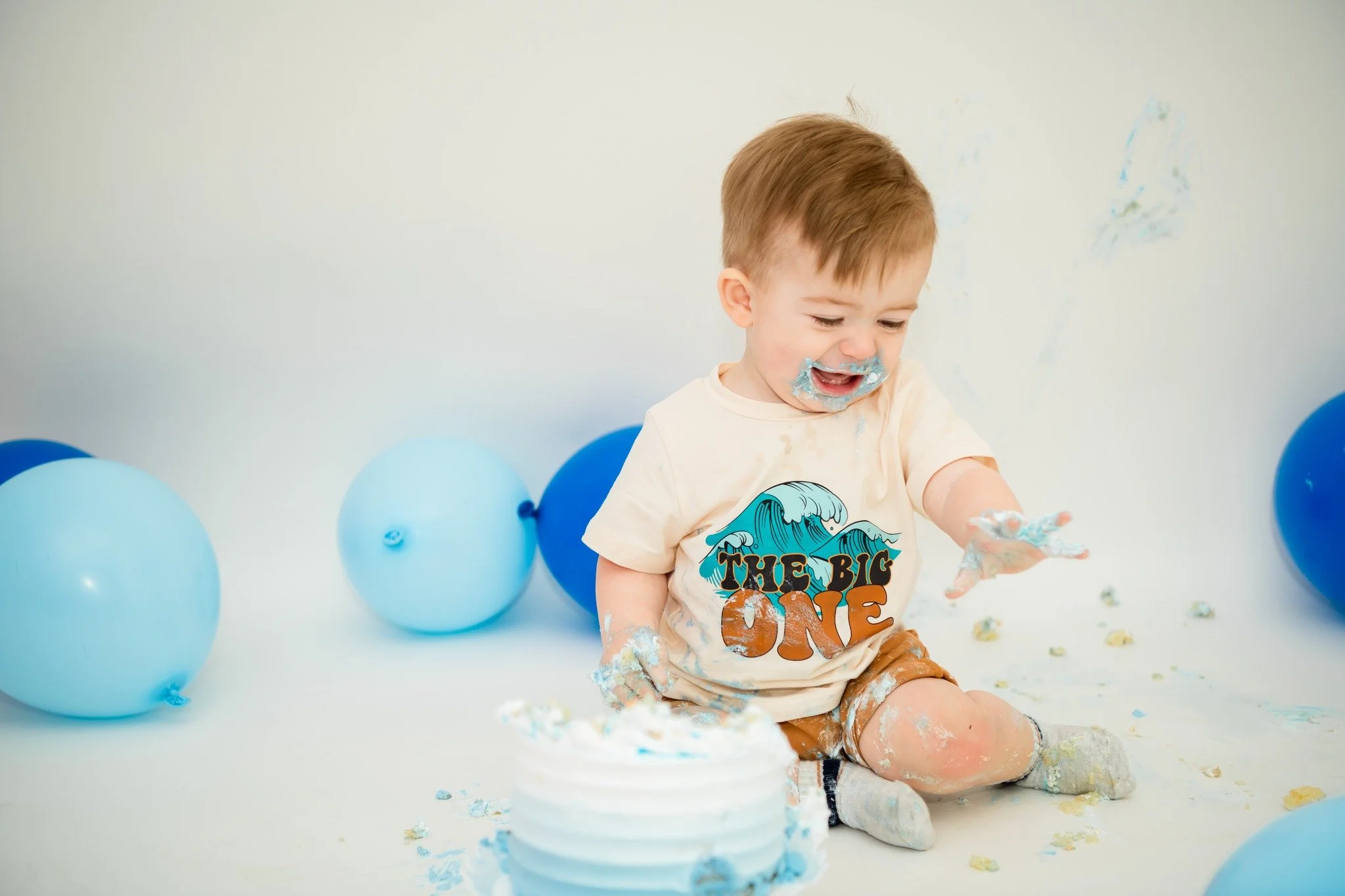 A young boy celebrating his first birthday, sitting on the floor with blue balloons around him, covered in cake and frosting, wearing a T-shirt that says 'The Big One'.