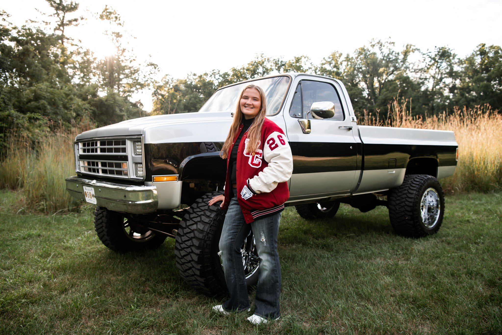 A young girl with long blonde hair wearing a red and white varsity jacket and jeans, smiling and standing next to a vintage black and white pickup truck on a grassy field with trees in the background during sunset.