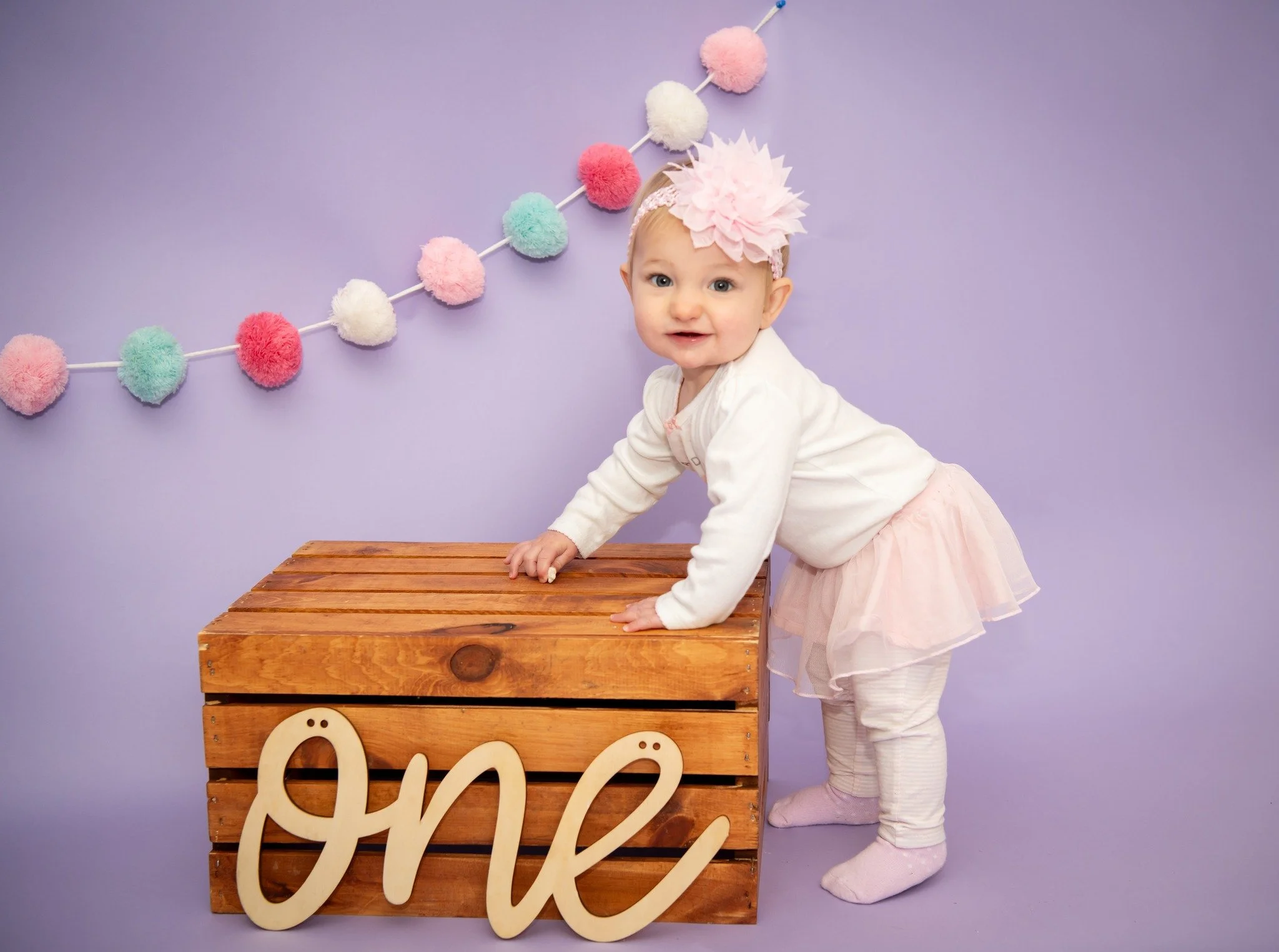 A baby girl dressed in a pink tutu, white cardigan, and pink headband with a large flower, leaning on a wooden crate with the word 'one' attached, against a purple background with a pastel pom-pom garland.