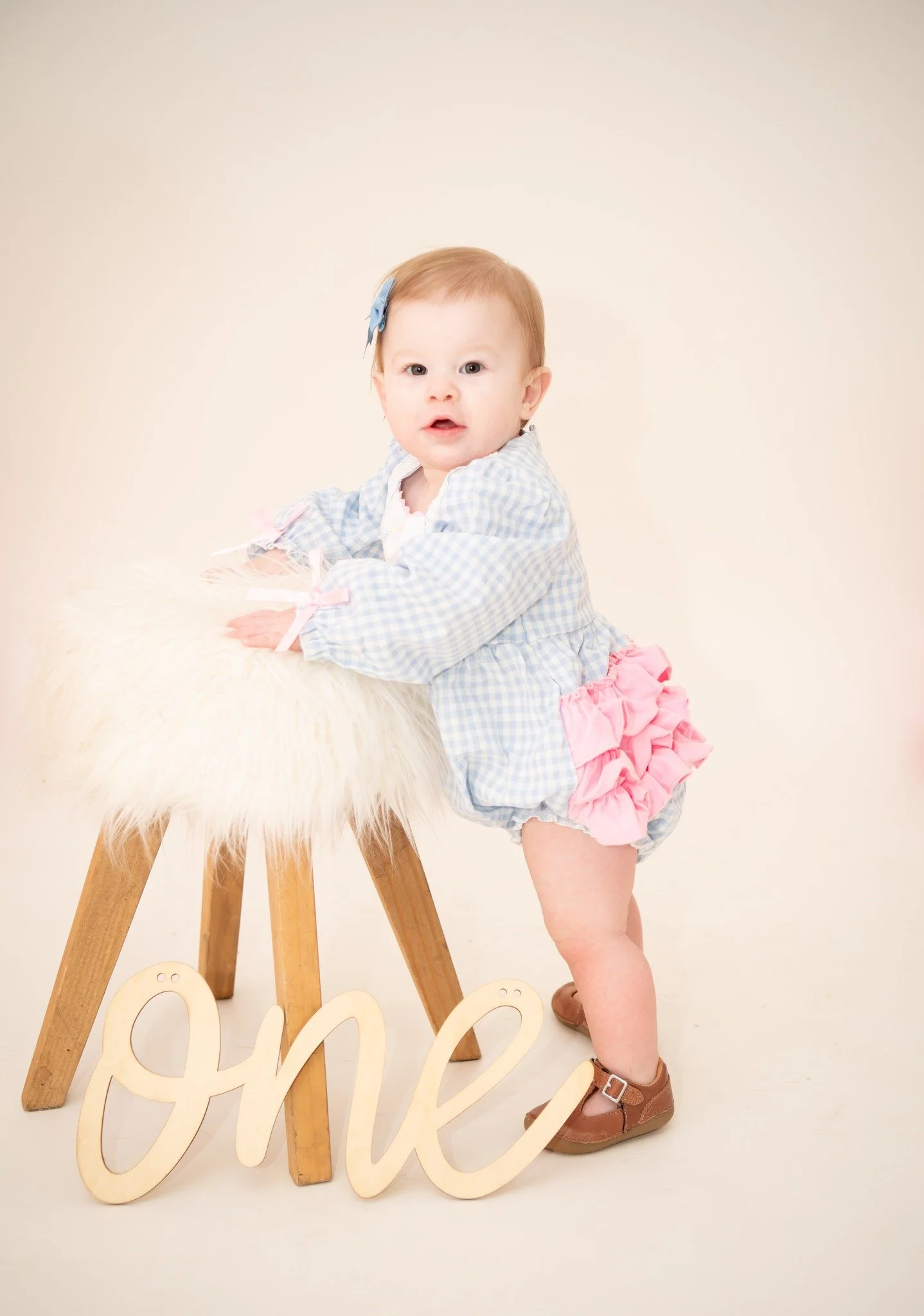 A young child with light hair, wearing a blue checkered top with pink ruffled shorts and brown shoes, stands next to a small wooden stool with a fluffy white covering, holding onto it. There is a decorative word 'one' in light-colored wood in front o