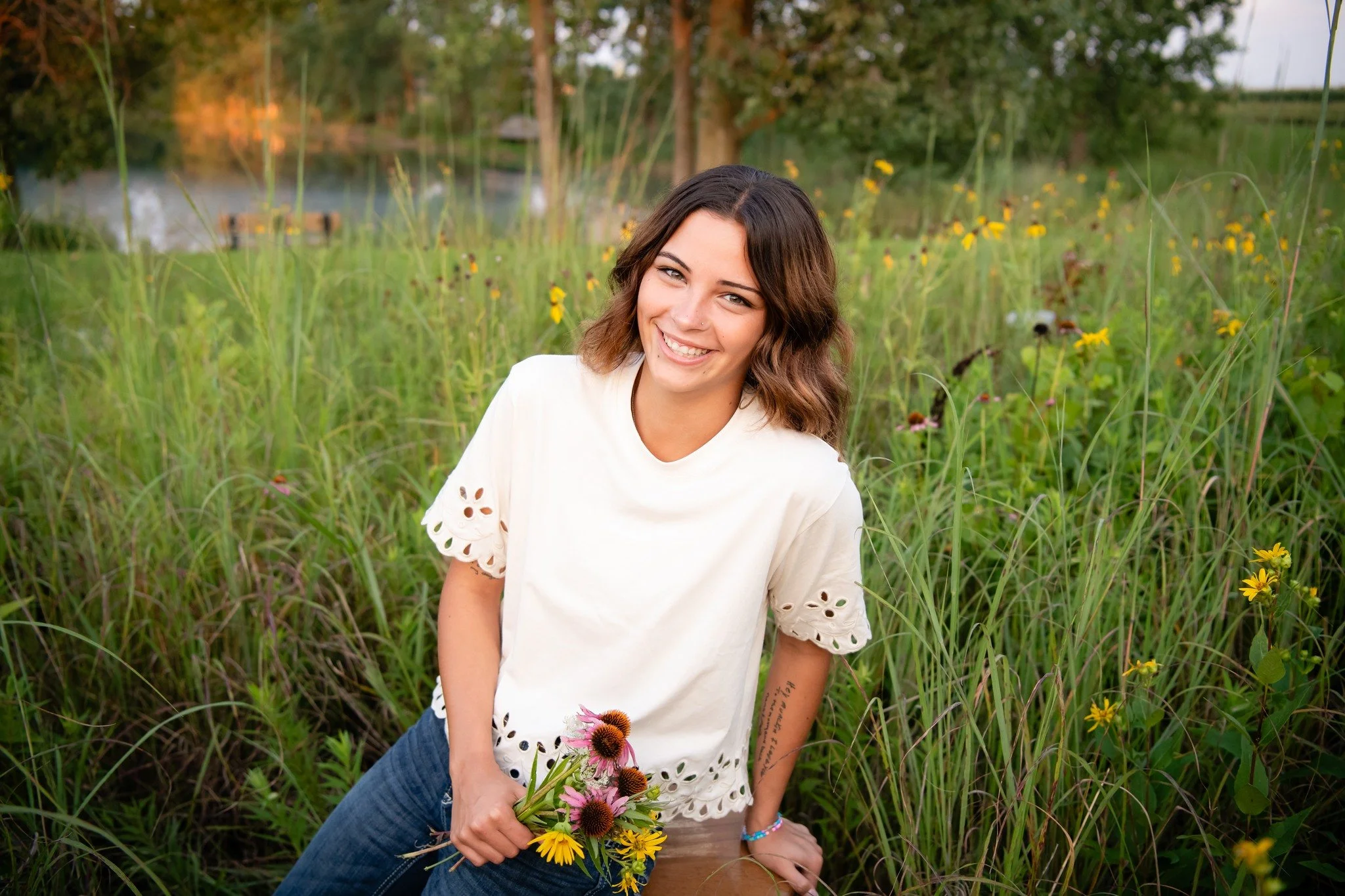A young woman with shoulder-length brown hair and a white top smiling while kneeling in a grassy field holding a bouquet of yellow and purple flowers.