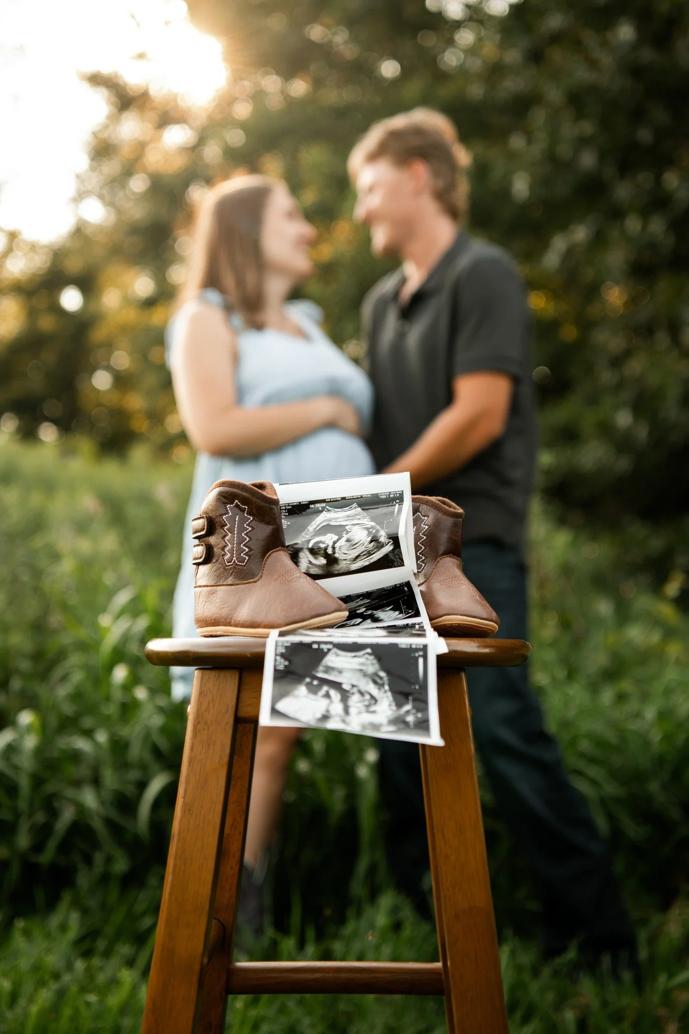 Baby shoes, ultrasound photos, and a couple in the background, with a pregnant woman, standing outdoors during sunset.