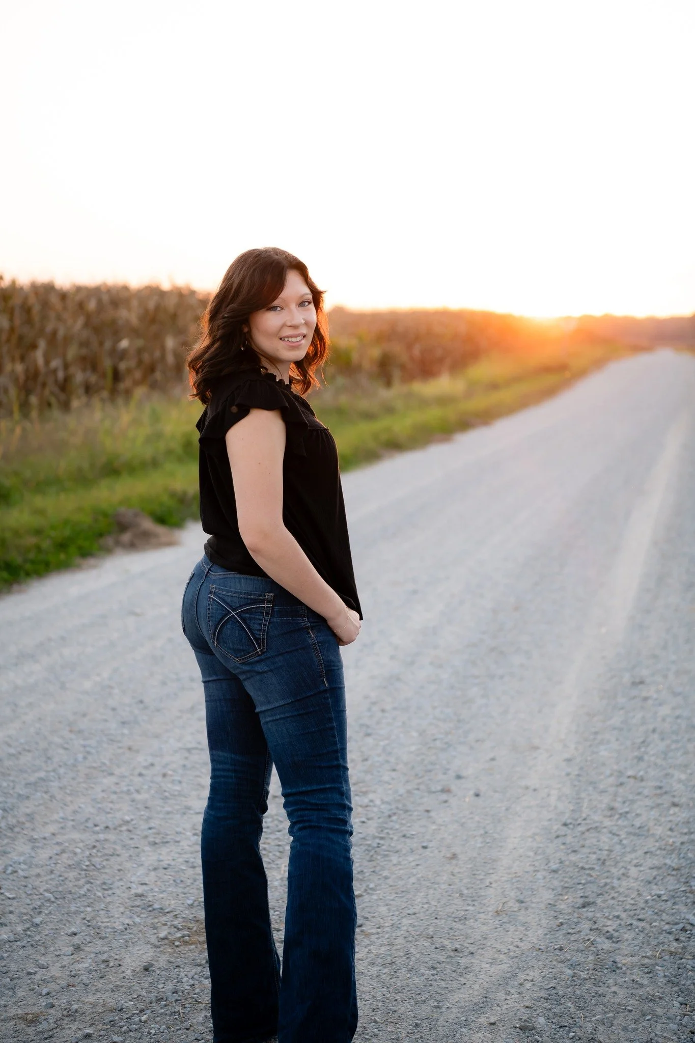 A woman with dark brown hair and a black top stands on a gravel road at sunset, with cornfields on each side.