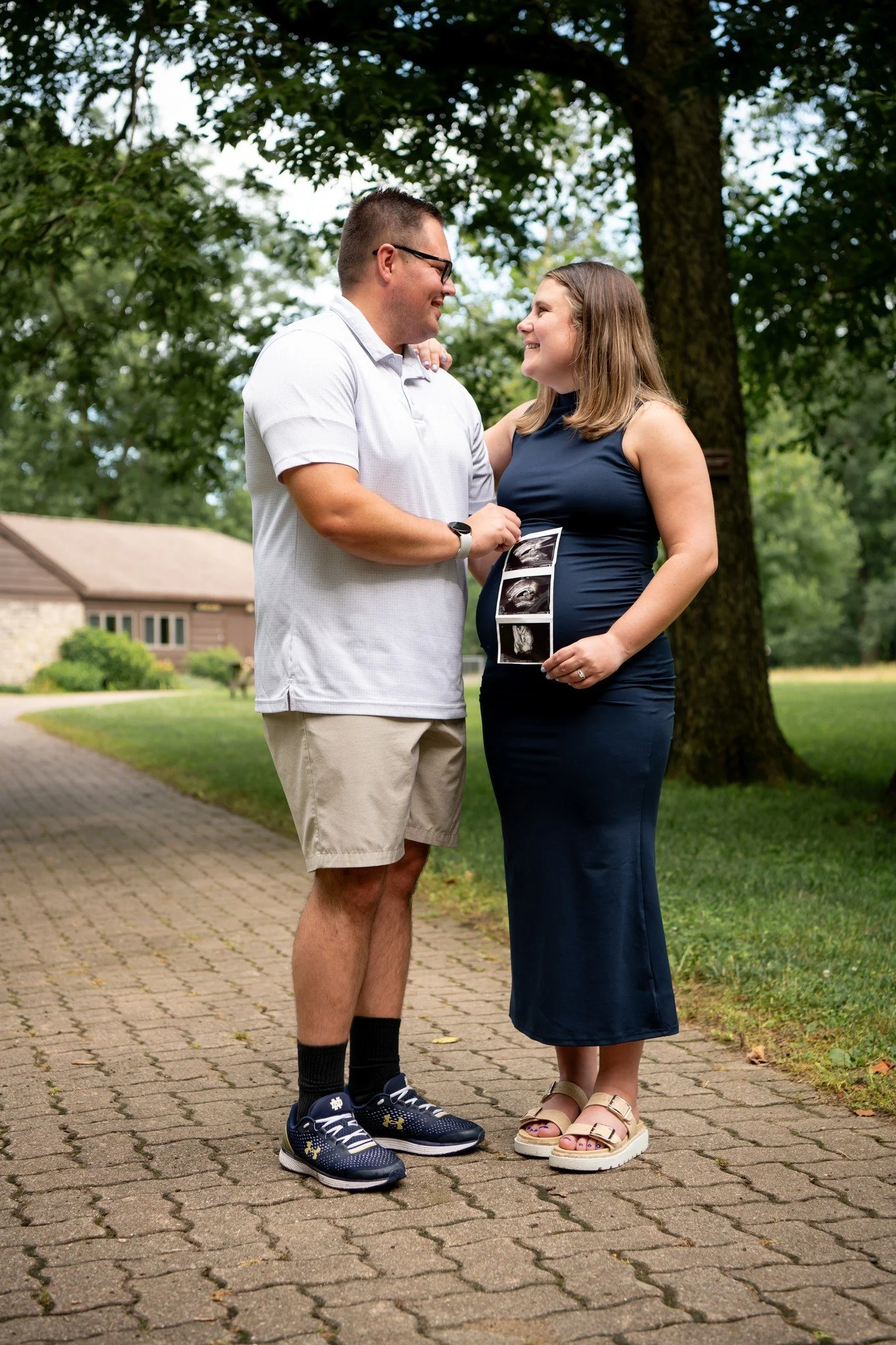 A couple standing outdoors on a brick walkway, with the woman showing her pregnant belly and ultrasound photos, and they are smiling at each other amidst greenery and large trees.