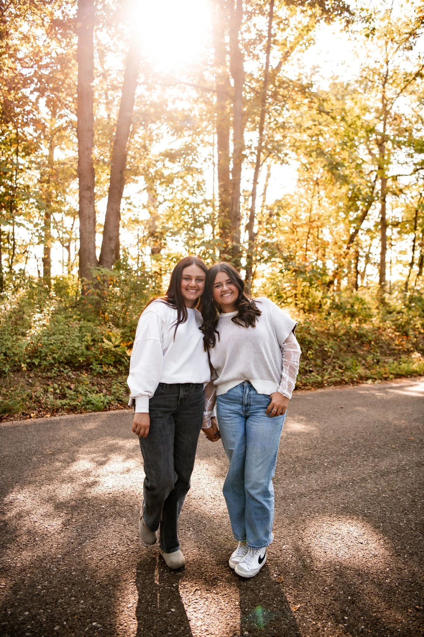 Two women stand together on a sunlit pathway in a forested area during fall. They are smiling and holding hands, dressed casually in white and light gray sweaters, with jeans and sneakers.