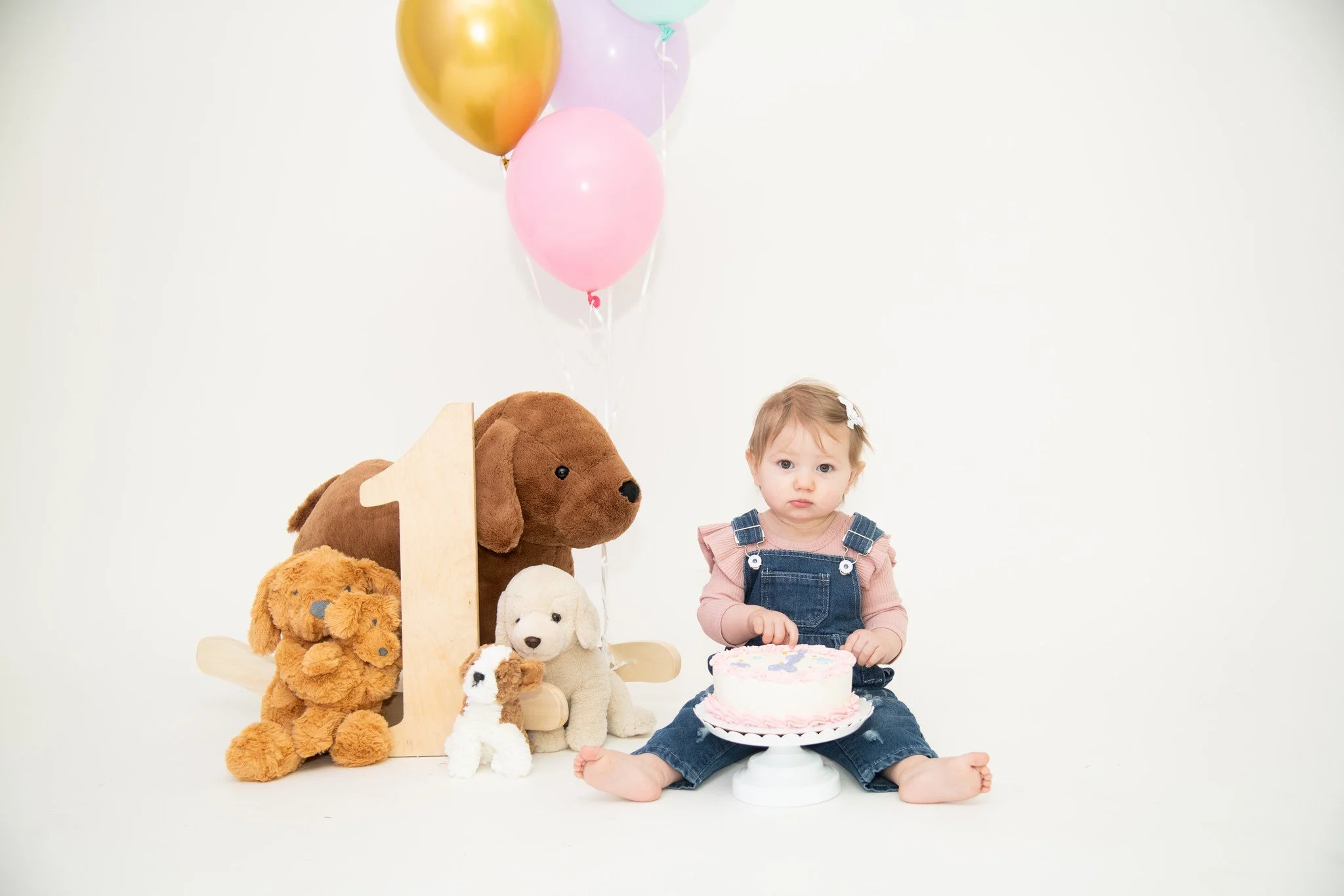 A toddler sitting on the floor with a birthday cake in front of her, surrounded by stuffed animals and balloons, celebrating her first birthday.