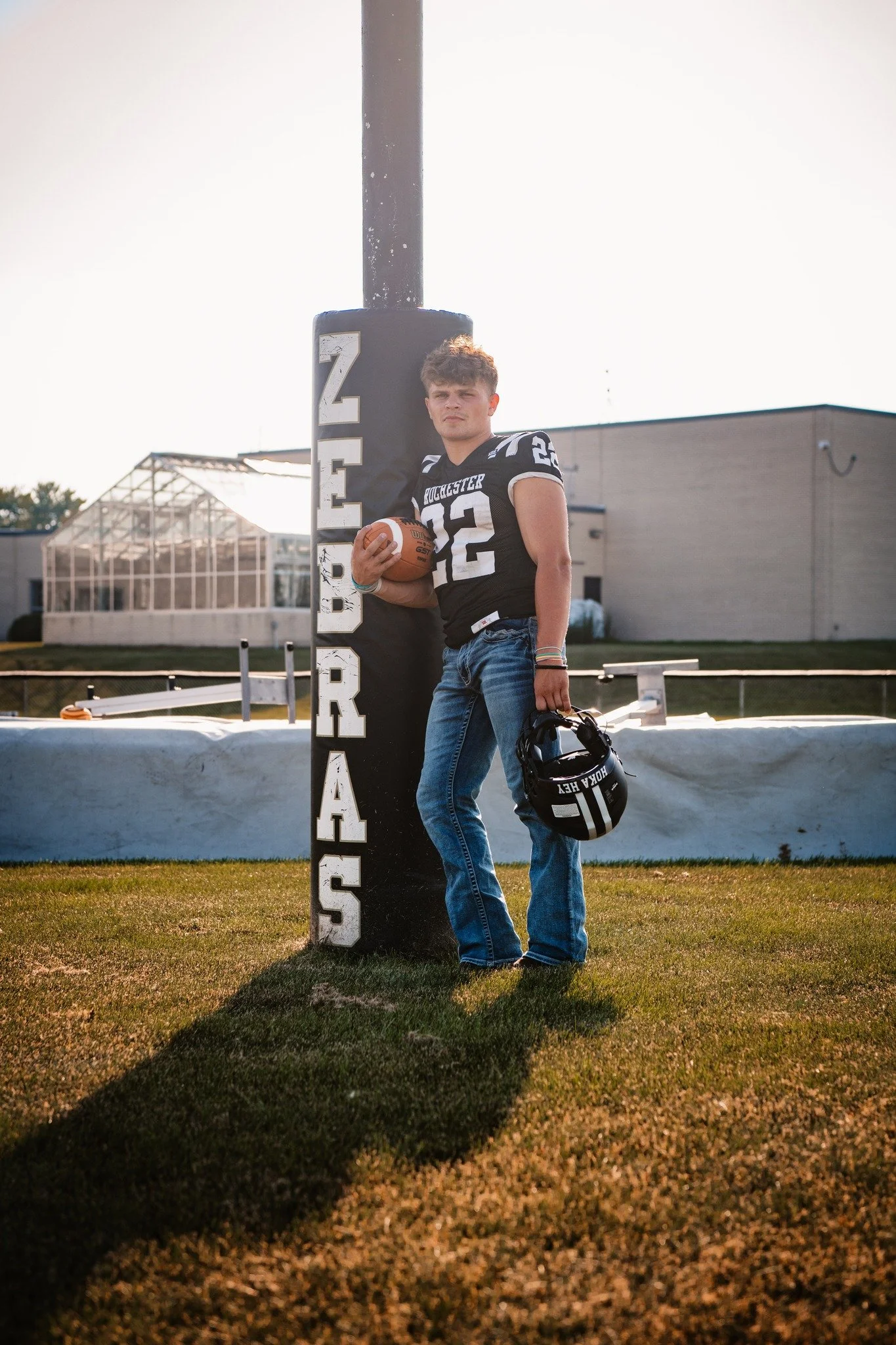 A teenage football player in a black jersey with the number 12 stands against a goalpost at a football field during the late afternoon. He is holding a football in one hand and a helmet in the other, with the word 'ROCHESTER' visible on his uniform a