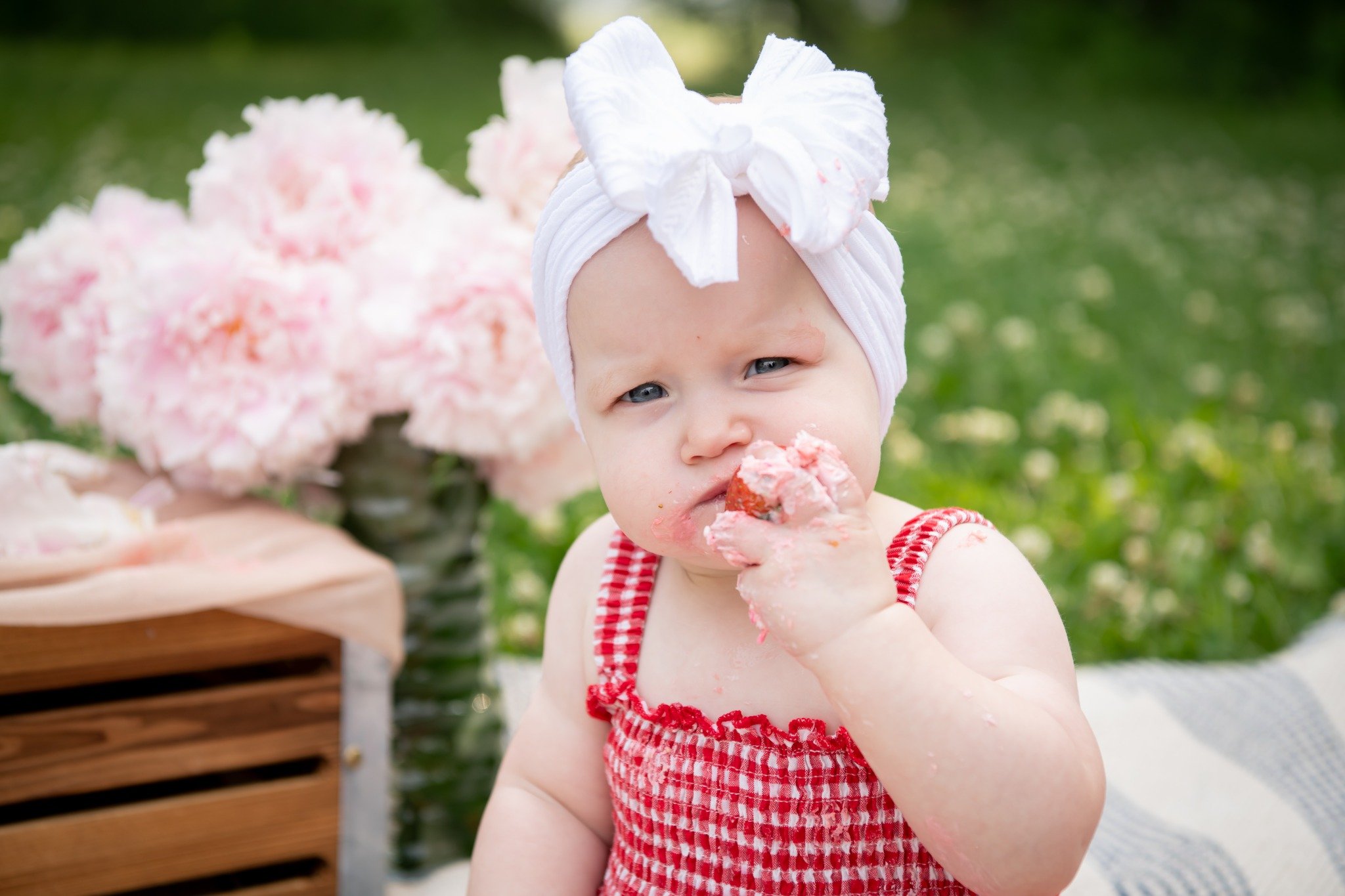 A baby girl wearing a white headwrap and a red gingham dress, eating a strawberry cake outdoors with pink flowers in the background.