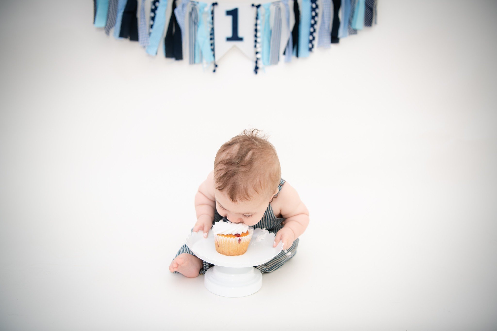 A baby sitting on the floor in front of a white background, leaning over a cake on a pedestal, with a blue and white birthday banner hanging above.