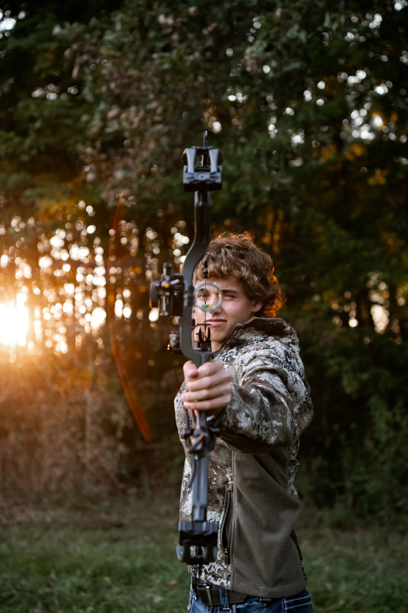A young man with curly hair holding a modern compound bow, aiming outdoors during sunset with trees in the background.