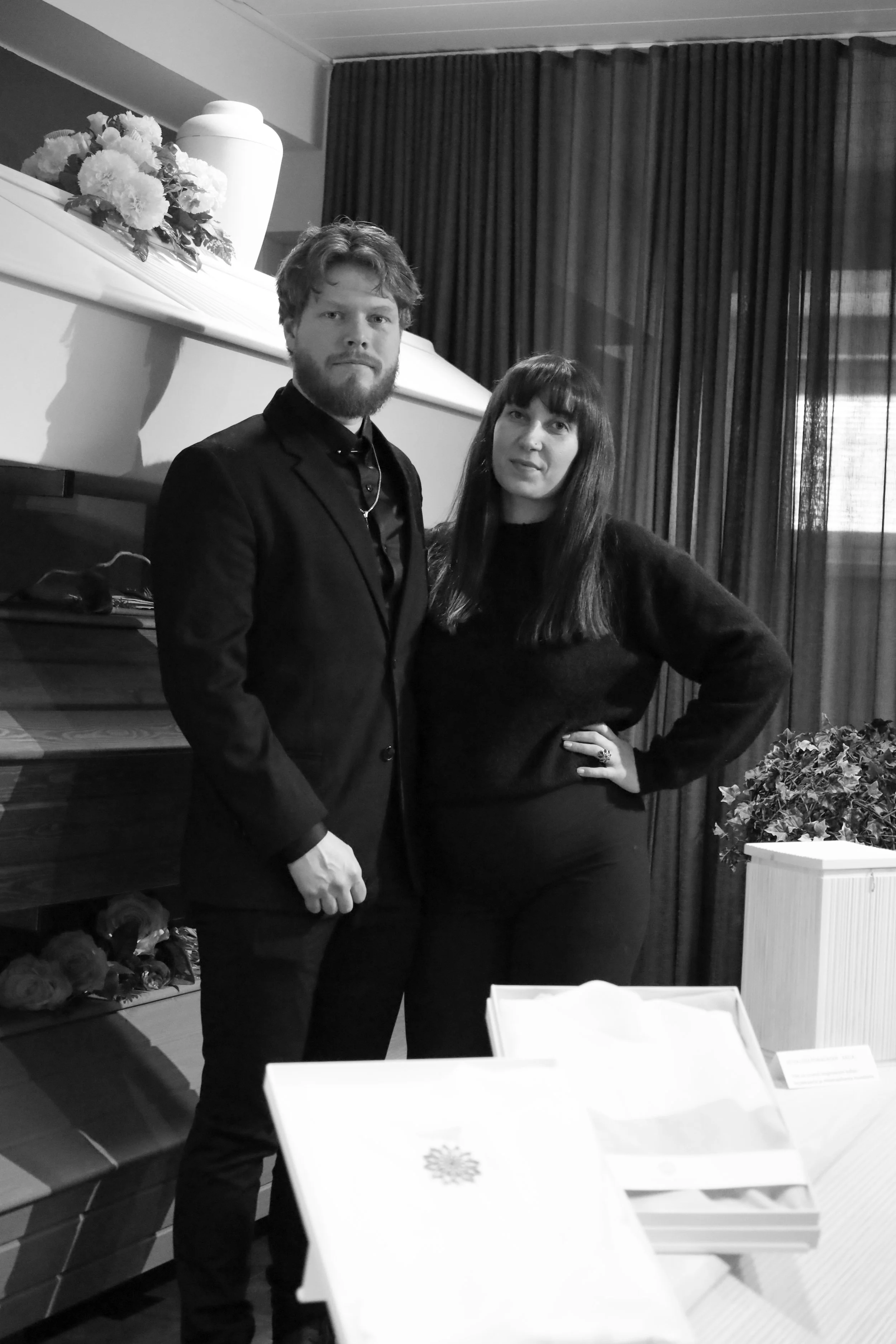 Kaksi ihmistä, mies ja nainen, seisovat sisällä, taustalla verhot ja sisustustavaroita, mustavalkokuva.