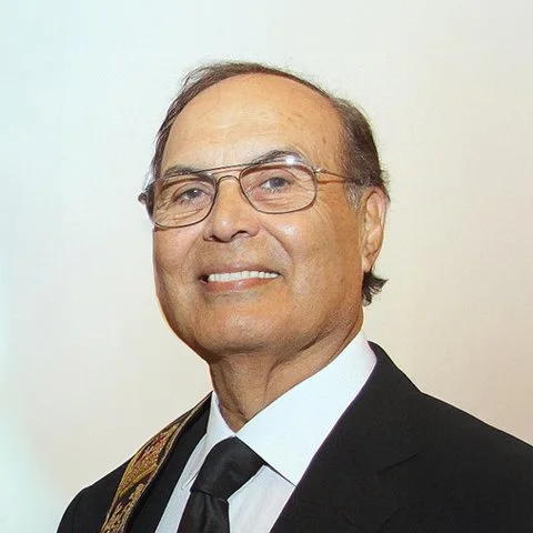Portrait of an older man wearing glasses, a black suit, white shirt, and black tie.