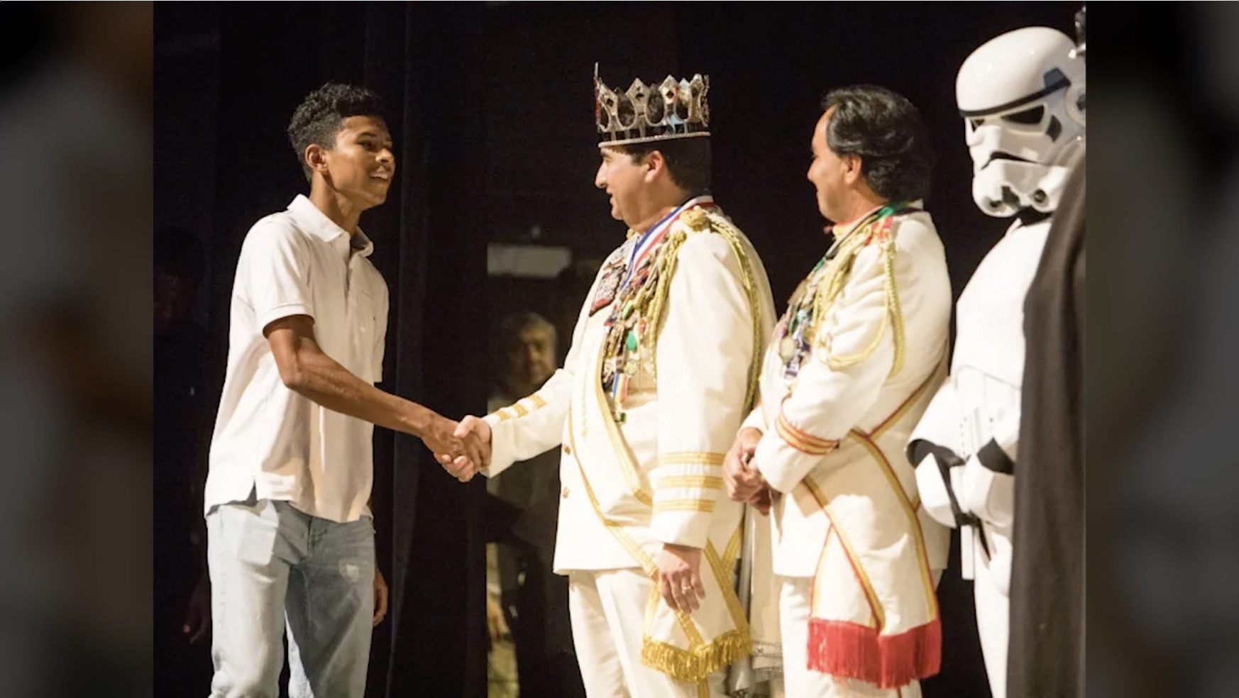 Young man shaking hands with a crowned man in a white and gold uniform, with two other similarly dressed men and a person in a Stormtrooper costume in the background.