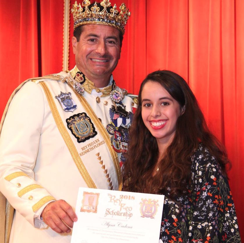 A man wearing a crown and a white military uniform adorned with medals and decorations, standing next to a young woman with long curly hair and a floral dress. The man is holding a certificate, and both are smiling in front of red curtains.