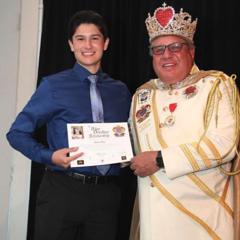 A young man in a blue shirt and silver tie holding a scholarship certificate, standing next to a man dressed as a king with a crown, royal robes, and medals, in front of a black curtain.