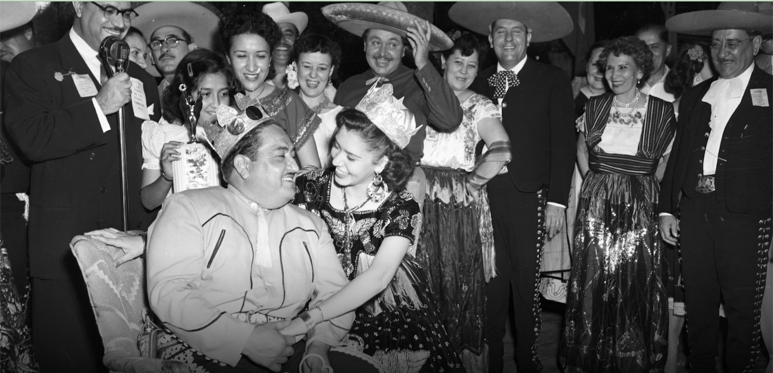 A group of people celebrating, with two in the center wearing crowns and traditional Mexican attire, smiling and looking happy, at what appears to be a festive event or fiesta.