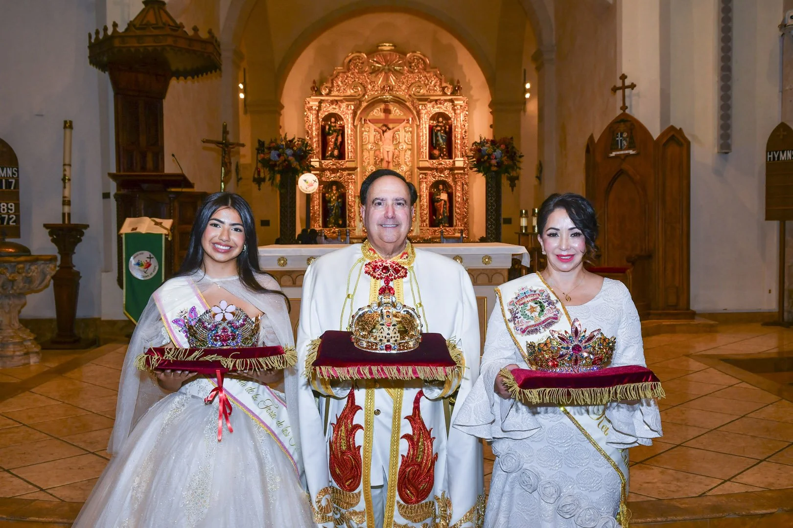 A man and two women standing inside a church, holding red velvet pillows with jewelry and crowns, dressed in traditional Filipino clothing for a fiesta celebration, with the church altar in the background.