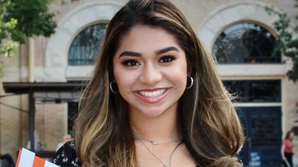 A young woman with long, wavy brown hair and hoop earrings, smiling outdoors in front of a building with large arched windows and trees.