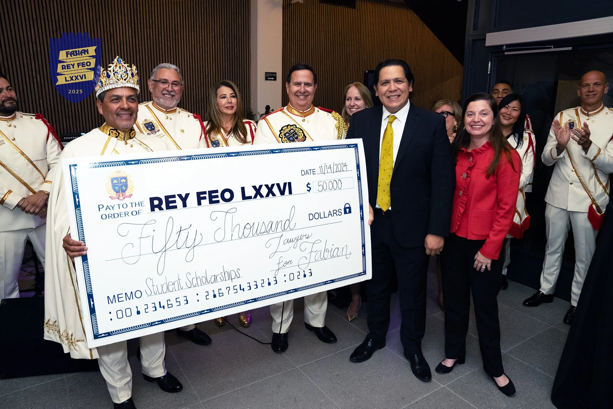 Group of people, including men in white ceremonial uniforms with gold trim and a crown, standing together holding a large check for fifty thousand dollars for student scholarships at an event celebrating Fabian Rey Feo LXXVI in 2025. Several people are smiling, and some are clapping.