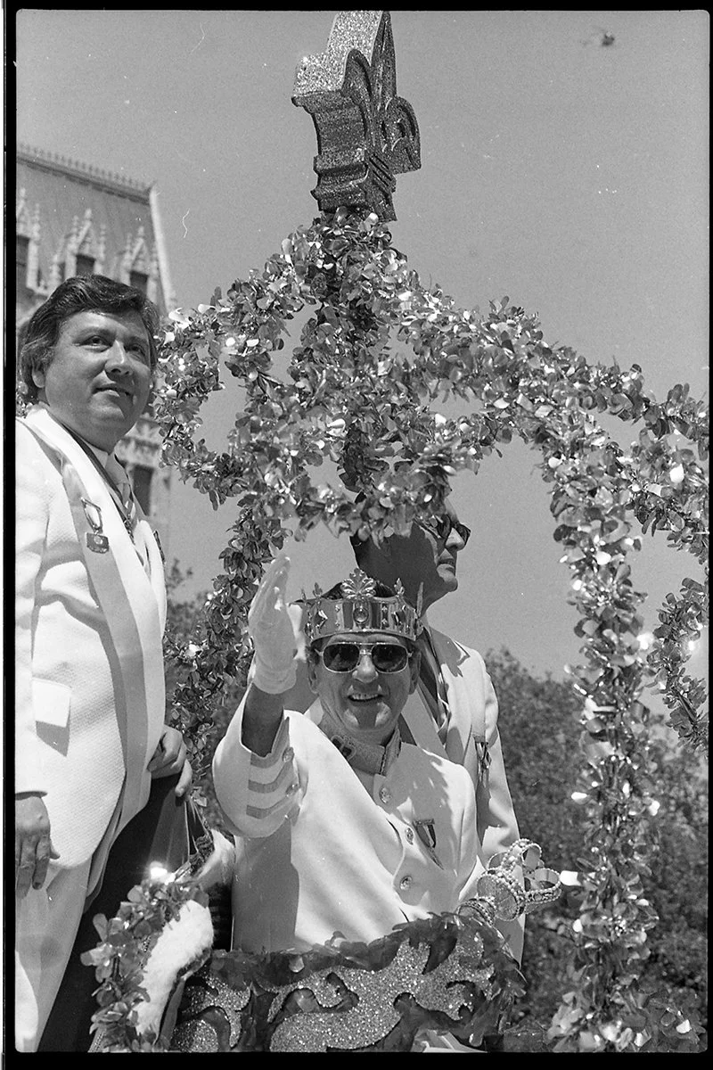 A woman wearing sunglasses and a crown is waving from a decorated float during a parade, with two men in light-colored suits standing nearby. The float is adorned with shiny garlands, and part of a building is visible in the background.