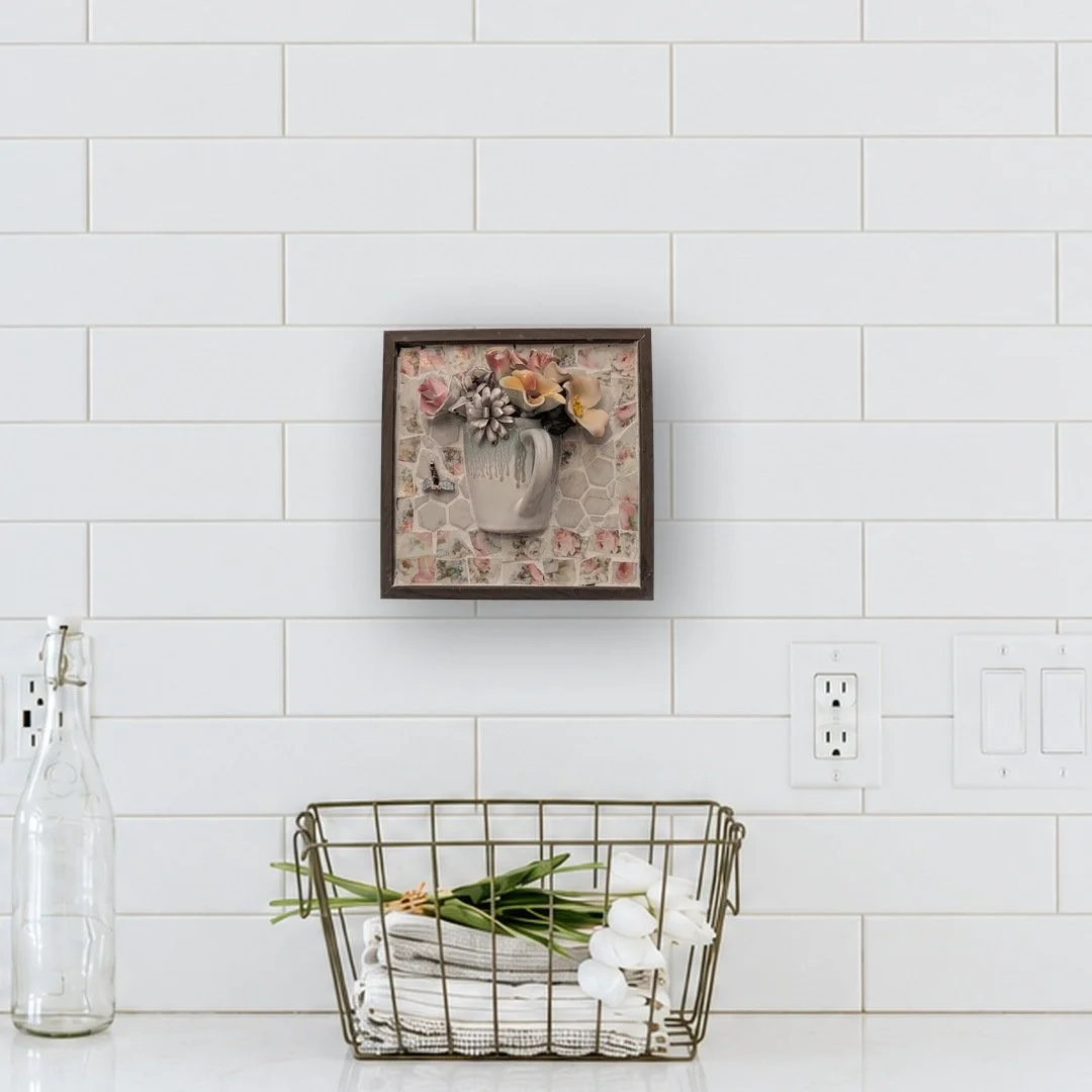Mosaic wall art of a framed half mug holding porcelain flowers, hung on a white tile kitchen wall above a wire basket