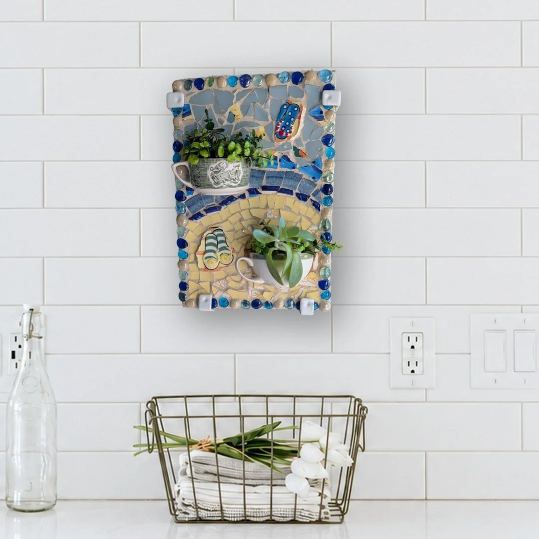 Mosaic wall art with two planted cups and flip-flop shards on blue and yellow china, hung on a white tile kitchen wall
