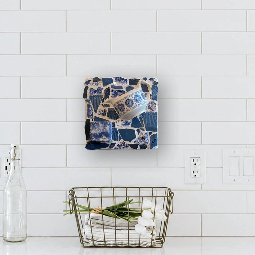 Blue and white mosaic wall art with a patterned cup on china shards, hung on a white subway tile kitchen wall