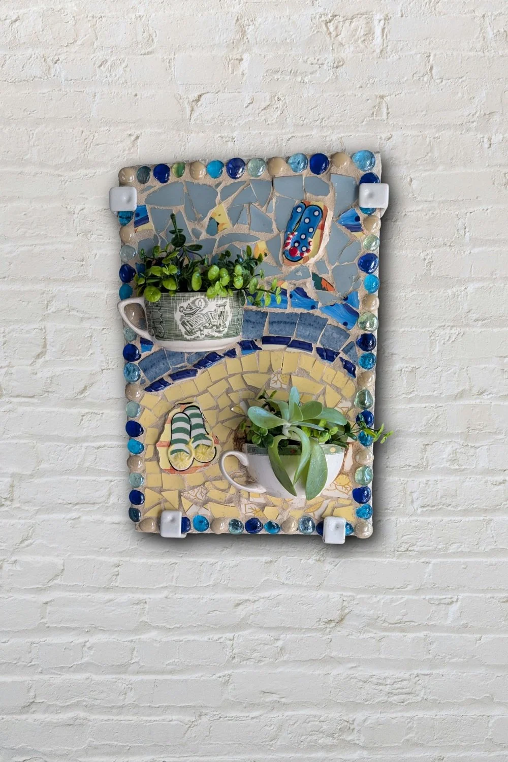 Cup of flowers and sandals mosaic wall art on white brick — outdoor-safe 3D assemblage by Sarasota artist Debra Rich
