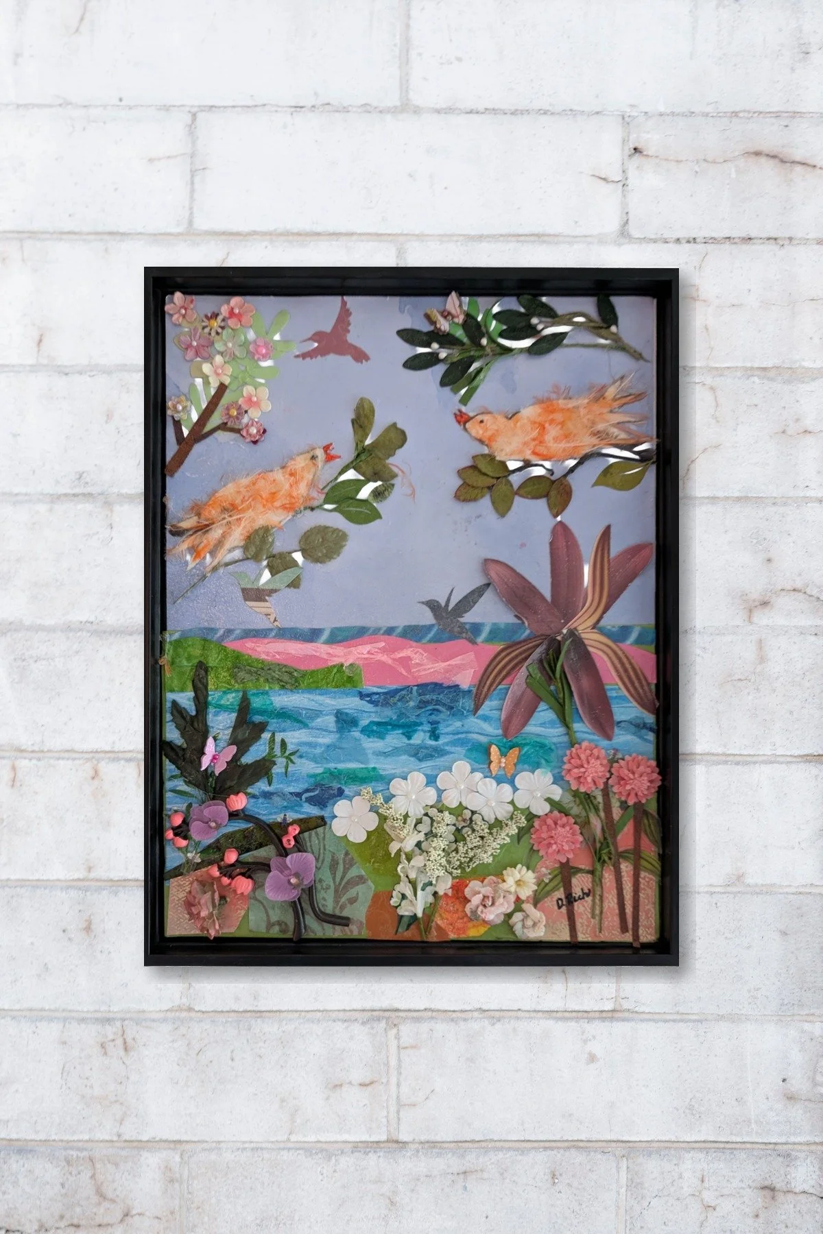 Mango's Last Stand framed mixed-media collage hanging on wall, two feather canaries in paper landscape with found-object flowers, by Debra Rich Sarasota -brick wall