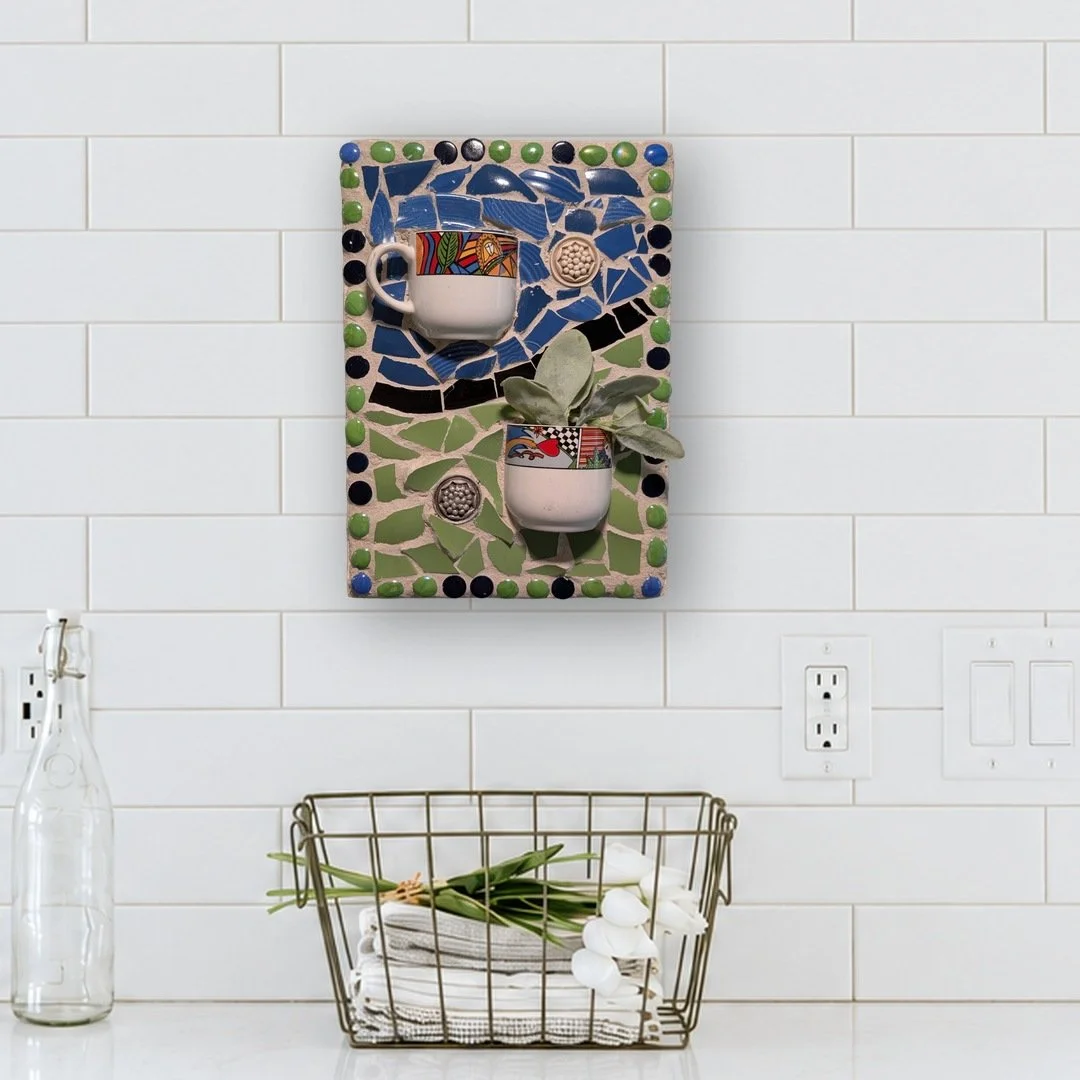 Tall mosaic wall art with two cups holding plants on blue and green china, hung on a white tile kitchen wall