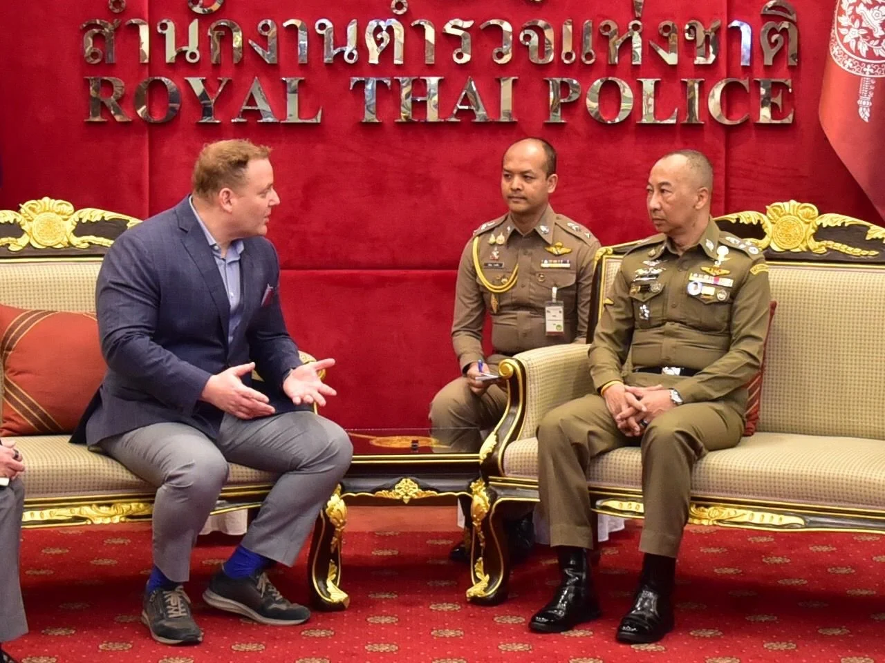 A man in a dark suit and light-colored pants is sitting on a sofa, engaged in conversation with two uniformed officers in front of a red wall with Thai and English signage reading 'Royal Thai Police'. The officers are dressed in khaki uniforms with m