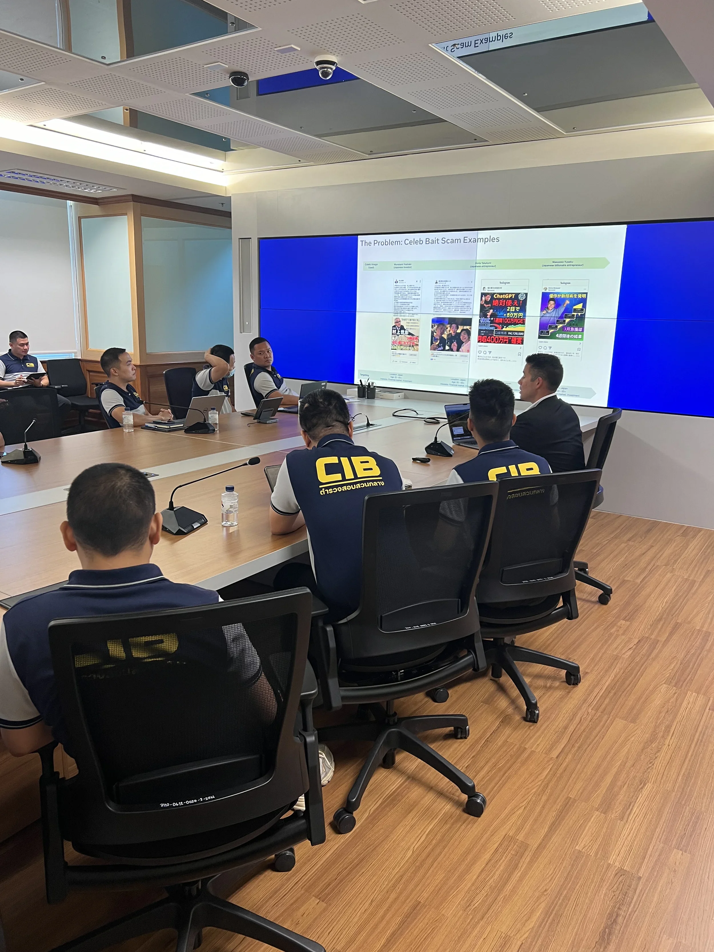 A group of people, some wearing uniforms with 'CIB' on the back, are sitting around a conference table in a modern office or meeting room, watching a presentation on a large screen displaying slides about scam examples.