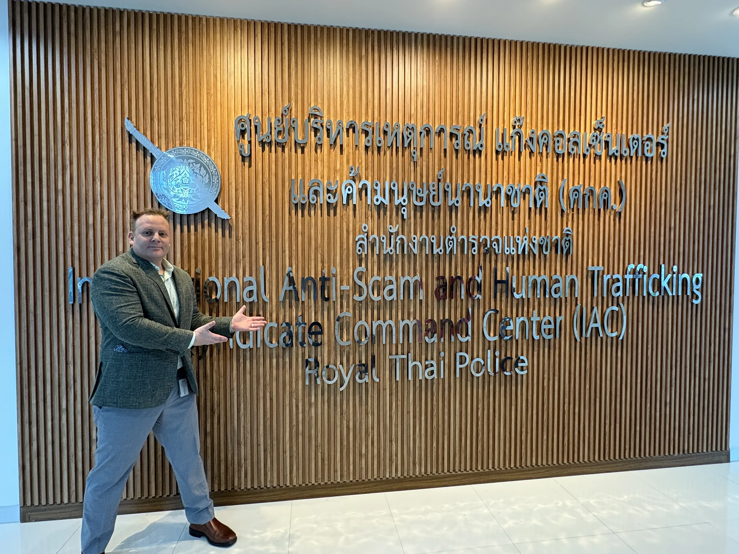 Man standing in front of a wooden wall with a sign for the International Anti-Scam and Human Trafficking Justice Command Center (JAC) at the Royal Thai Police, pointing towards the sign with his hands.