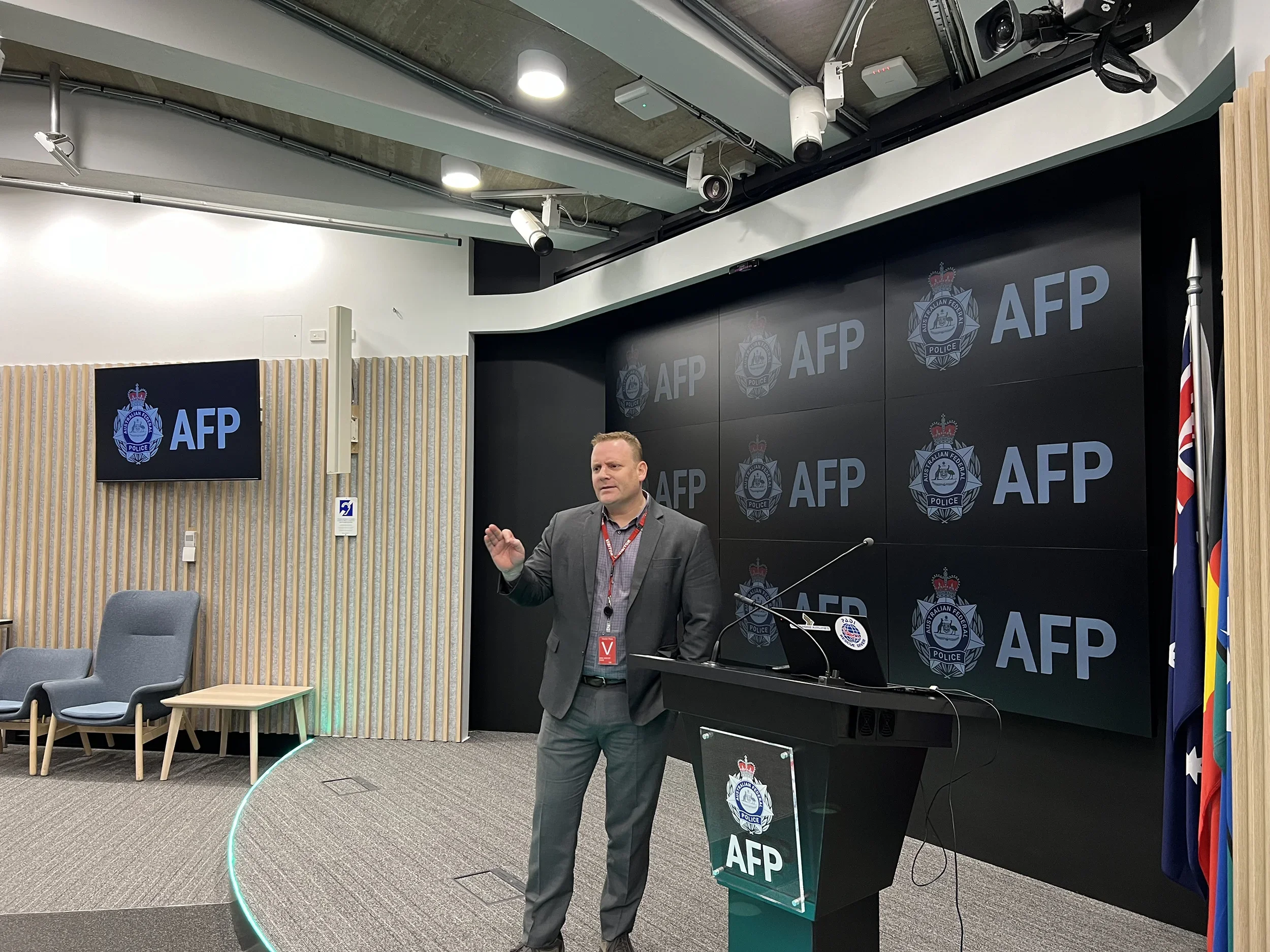 A man in a suit standing at a podium in a press conference room with police and Australian Federal Police (AFP) logos on screen behind him. Flags are visible on the right side.