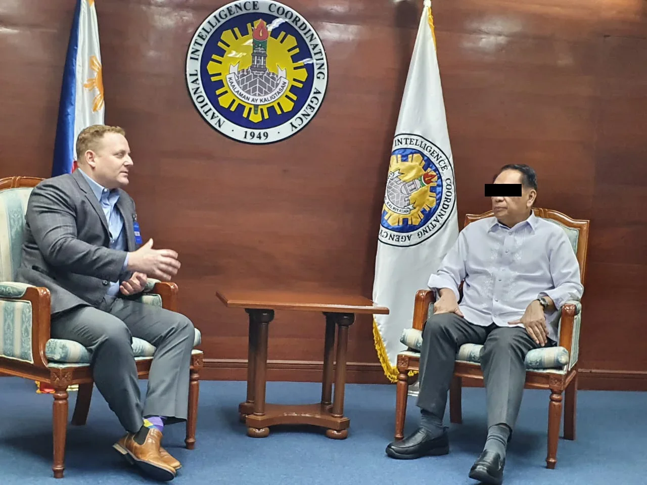 Two men are sitting in a formal room with wooden walls and flags. One man is speaking and gesturing with his hand. The man on the right is listening. There is a banner in the background with an emblem and the words 'National Intelligence Coordinating